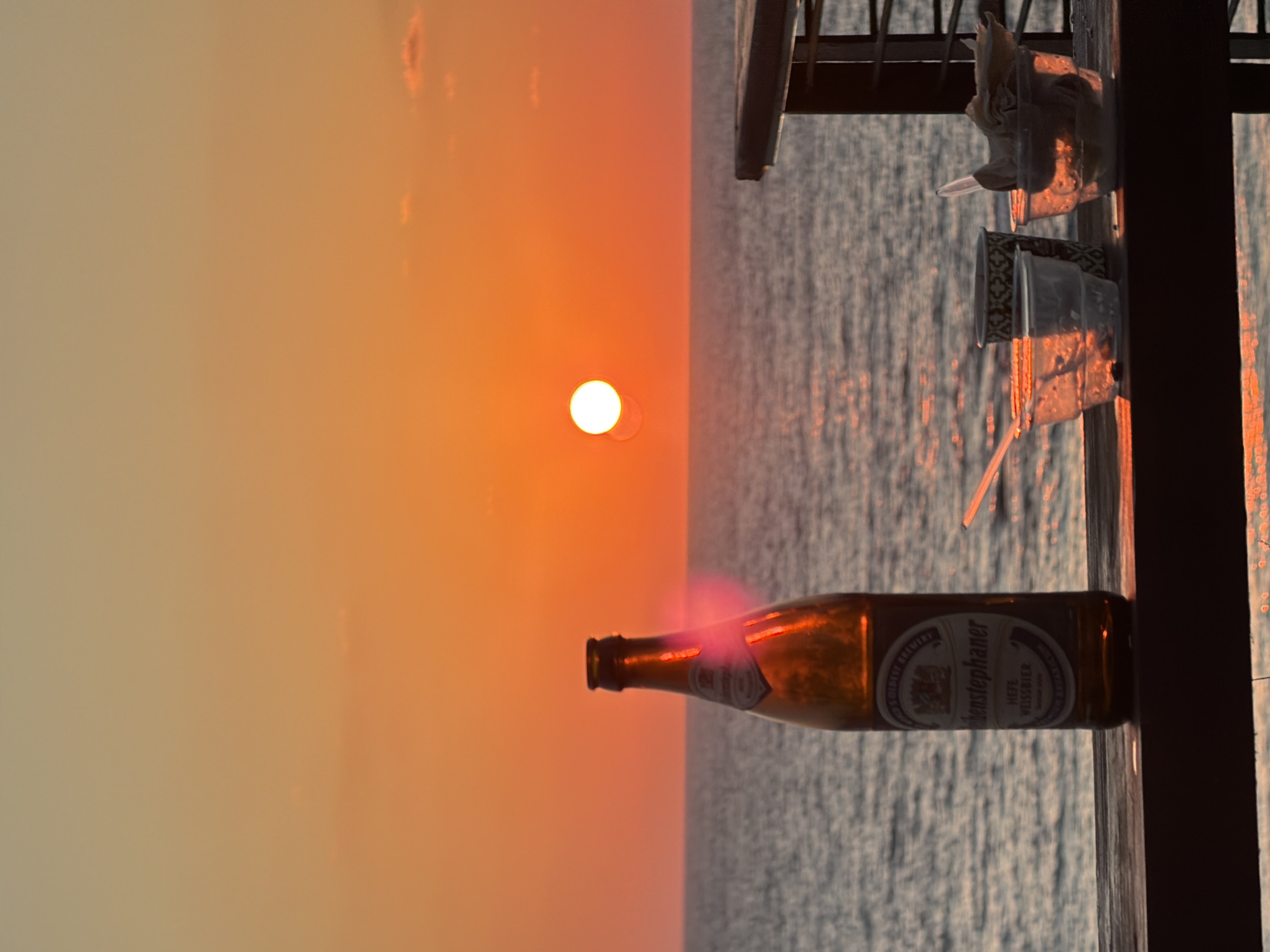 This image depicts a sunset over the Mediterranean Sea, viewed from a balcony or deck in Bat Yam, Israel. The sun is a bright, round orb positioned near the horizon, casting a warm orange glow across the sky and the water. In the foreground, a brown glass bottle of beer, identified by its label as "Benediktiner Hefe Weissbier," stands on a wooden railing. To the right of the bottle are several plastic cups, some appearing to contain remnants of food or drink, and a paper cup with a decorative pattern. The sea is calm, with gentle waves reflecting the sunset's light. The overall atmosphere is one of relaxation and appreciation of the natural beauty.