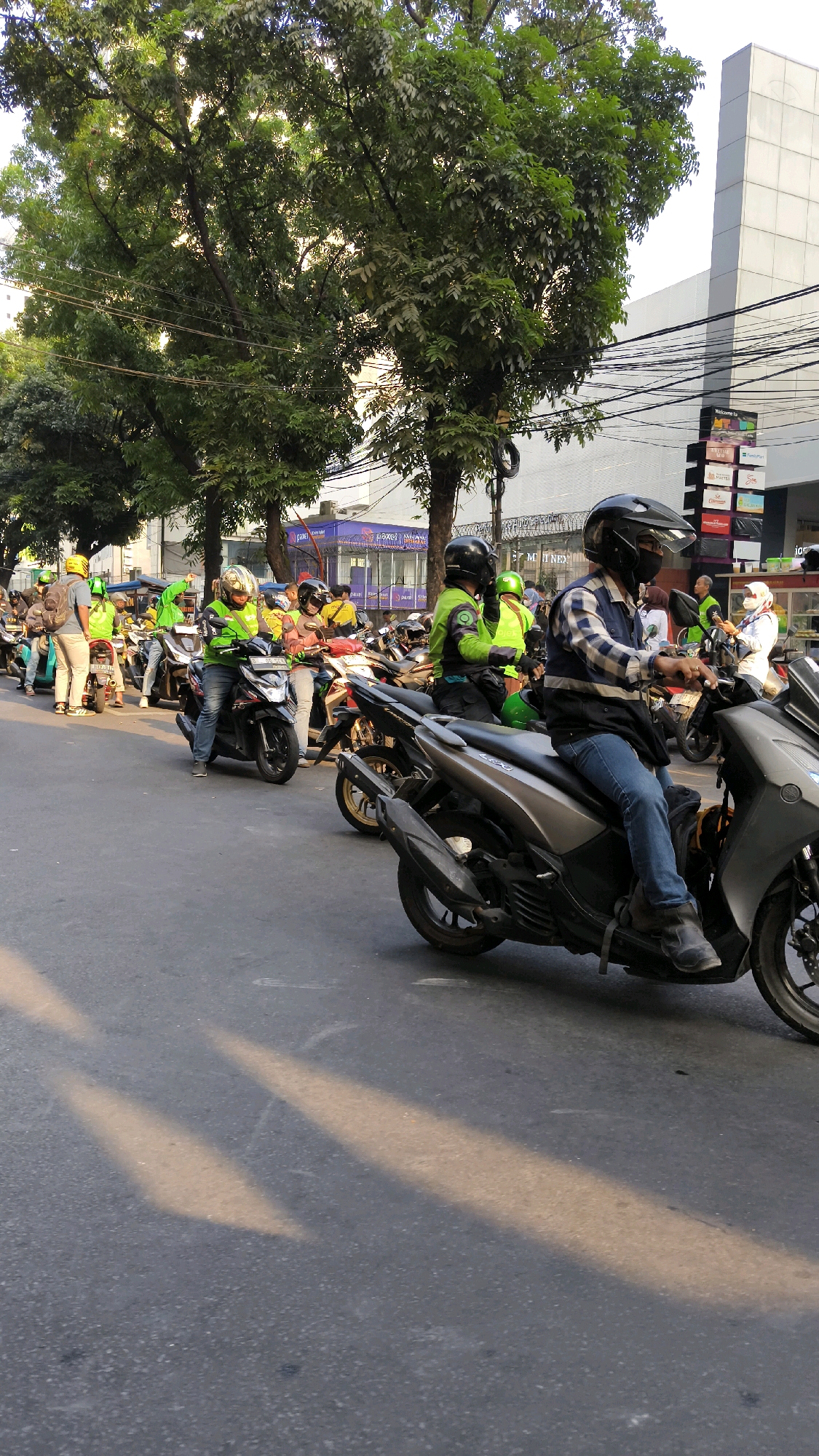 The image depicts a street scene in South Jakarta, Indonesia, characterized by a line of motorcyclists, many wearing bright green jackets, indicative of ride-sharing services. The motorcyclists are positioned on a paved road, with some stationary and others in motion, suggesting a traffic situation or a gathering.
In the foreground, a motorcyclist in a checkered shirt and dark vest is riding a silver scooter. His face is partially obscured by a black helmet and a black face mask. The scooter has a black windshield and silver accents. Behind him, another motorcyclist in a green jacket and helmet is visible.
Further down the street, a larger group of motorcyclists are lined up. Many are wearing similar green jackets and yellow or black helmets, with some also wearing backpacks. The motorcycles are a mix of models and colors, with a few prominent silver and black scooters.
The environment includes lush green trees lining the side of the road, with their branches reaching towards the sky. In the background, a modern building with a white facade stands tall, interspersed with power lines and street signs. The lighting suggests it is either early morning or late afternoon, with sunlight casting distinct shadows on the road. The overall scene conveys a sense of urban mobility and the presence of a significant number of motorcycle couriers.
