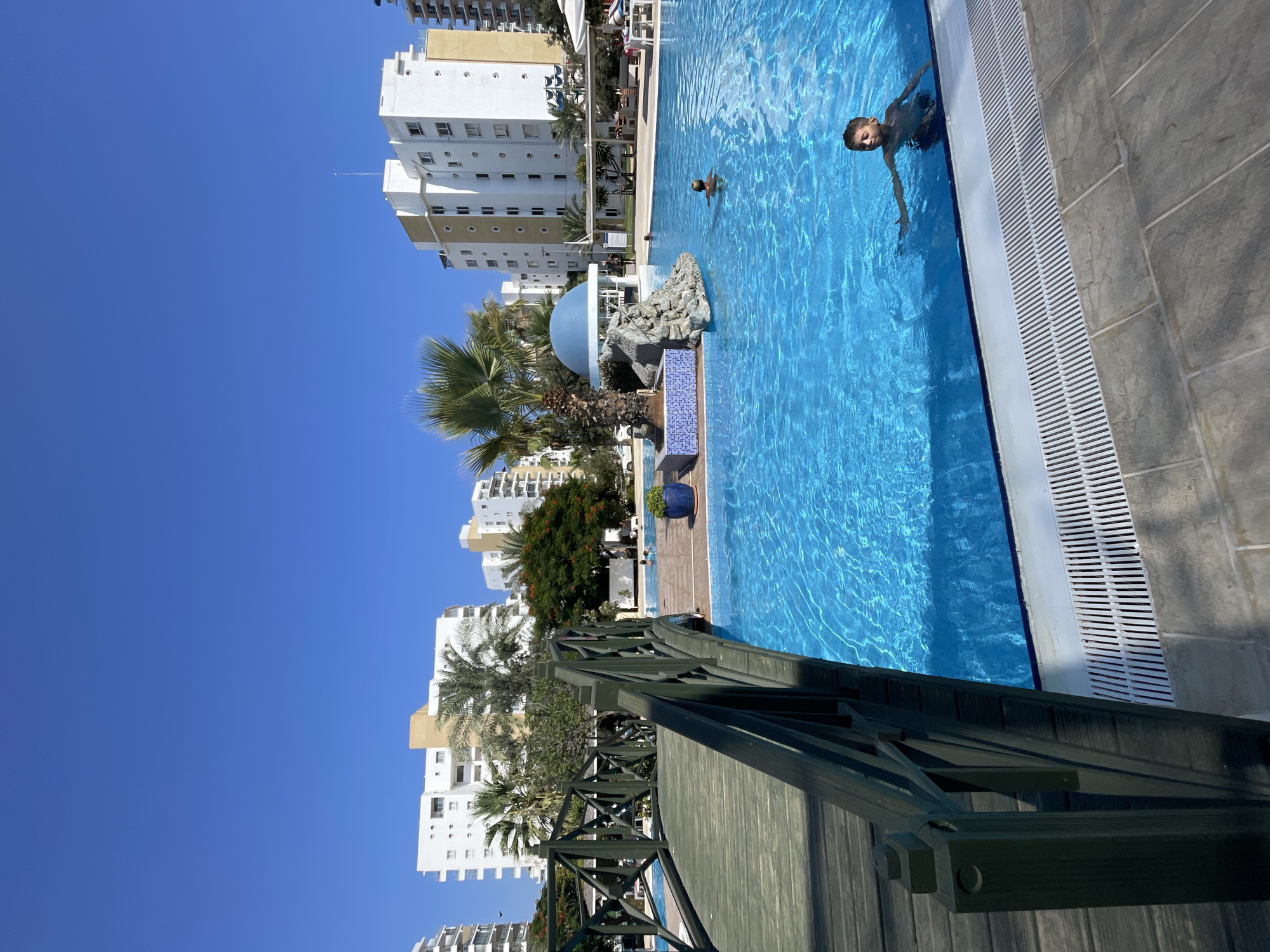 The image depicts a sunny outdoor scene at a resort or residential complex, likely in Trikomo, Northern Cyprus. A large, bright blue swimming pool occupies the foreground and midground, with crystal clear water reflecting the clear blue sky. A dark green wooden footbridge with intricate railings extends into the frame from the left, arching over the pool.
In the pool, two young boys are visible. One boy, positioned closer to the foreground and right of the frame, is partially submerged in the water, looking towards the camera with his arm outstretched. Another boy is further back in the pool, appearing to swim.
In the background, several multi-story white and beige buildings with balconies are visible, indicative of a Mediterranean architectural style. Palm trees and other lush greenery are scattered around the buildings and the pool area, suggesting a warm climate. To the left of the pool, a small, decorative island features a blue domed structure resembling a gazebo, surrounded by rocks and potted plants. Other elements of the scene include lounge chairs and tables, some covered by white umbrellas, hinting at relaxation and recreational areas. The overall impression is one of a serene and idyllic vacation or residential setting.
