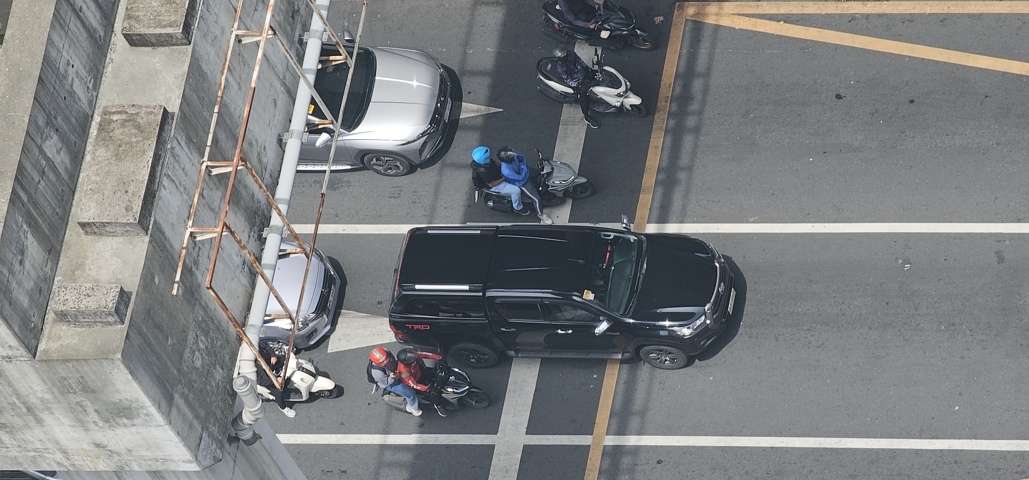 This aerial image, taken in Makati, Philippines, captures a traffic scene on a multi-lane road. In the foreground, a black pickup truck is parked and angled slightly. To its left, a silver car is also stopped, with a white scooter parked beside it, occupied by a person in dark clothing and white sneakers. Another scooter, occupied by two individuals wearing red and blue jackets and helmets, is positioned behind the silver car and in front of the black pickup truck.
Further up the road, a silver car is stopped, and to its right, a scooter with two passengers wearing blue and black is visible. Above this, another scooter is parked with a person in a patterned jacket and black pants. The furthest visible scooter has a rider in a dark jacket and pants. The road surface is asphalt with white and yellow lane markings. The scene is bordered on the left by a concrete structure with vertical metal supports, suggesting an elevated road or bridge. The lighting indicates it is daytime, with shadows cast by the vehicles and the overpass.