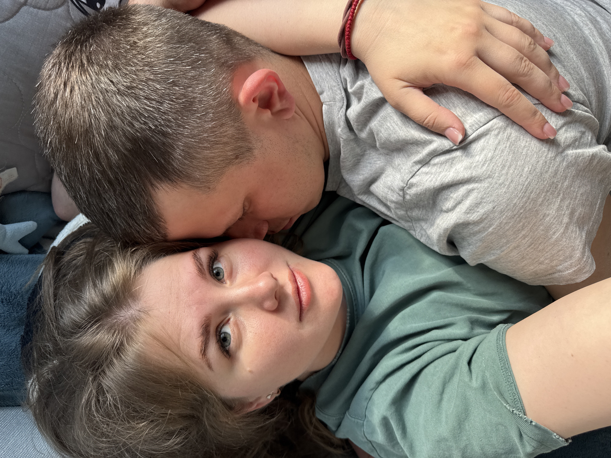 A close-up, elevated shot depicts a couple lying down. The woman is facing the camera, her gaze directed at the viewer. She has fair skin, light brown hair, and blue eyes. Her clothing is a green t-shirt or hoodie. A man is lying behind her, with his face turned towards her cheek. He has short, dark hair, and his body is covered with a light grey t-shirt. His hand rests on the woman's shoulder, with a red bracelet visible on the wrist. The location appears to be indoors, likely a home setting, as suggested by the casual attire and relaxed poses. The exact city of Ферма, Russia is impossible to infer with just this photograph.