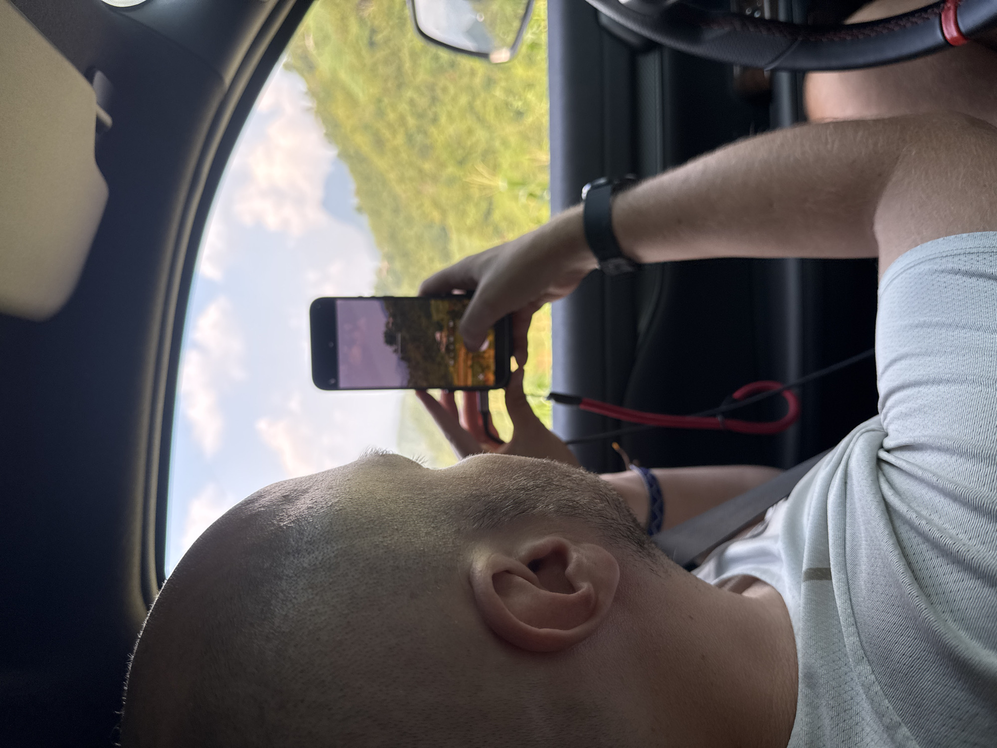 A man is seated in a car, holding a smartphone in his hand, taking a picture or video of the scenery outside the window. The exterior view is of green vegetation, possibly trees, under a sky with some clouds. The interior shows the car's steering wheel, part of the dashboard, and the side mirror. The man is wearing a white shirt and a seatbelt. A red cable is visible near his arm. The location could be Mậu Duệ Commune, Vietnam, based on external information, as this detail is not visually discernible from the image alone.