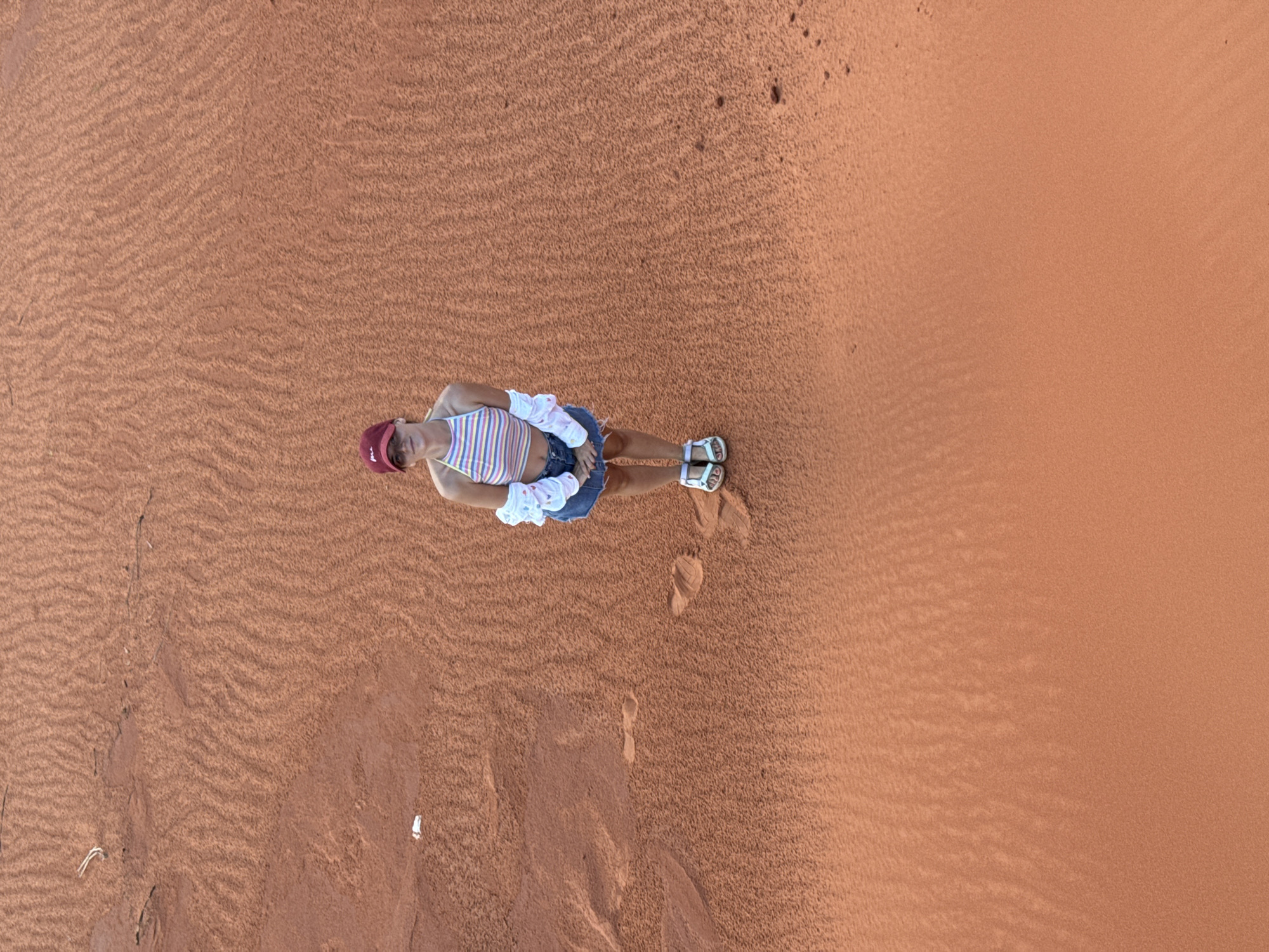 An aerial, eye-level shot shows a person standing in the sand dunes of Mũi Né, Vietnam. The person is a female wearing a red baseball cap, a multi-colored striped halter top, a white unbuttoned shirt, and a blue denim skirt. She is wearing white sandals. The sand dunes are reddish-orange with rippling patterns from the wind. There are a few footprints visible in the sand.