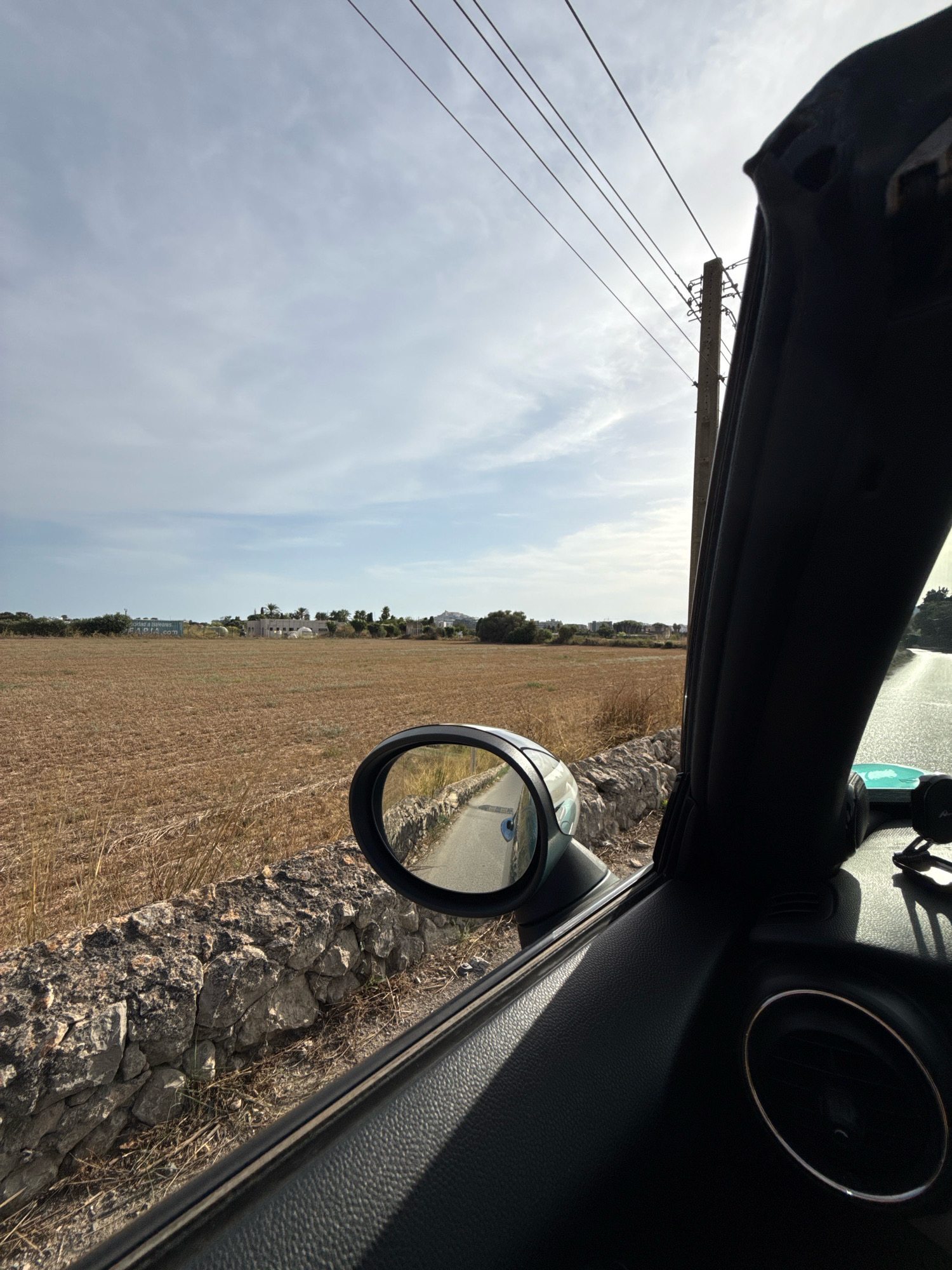 The image depicts a view from inside a vehicle looking outwards, on a road likely in Santa Eulària des Riu, Spain. The foreground shows the interior of the car, including a black dashboard and a side mirror. The side mirror reflects a narrow road, a stone wall, and dry, brown vegetation. Beyond the road and wall, a large field of dry, stubbly crops stretches towards a distant cluster of buildings. The sky is partly cloudy, with a utility pole and overhead wires visible against it. The scene suggests a rural or semi-rural setting, possibly during the late summer or autumn harvest season.