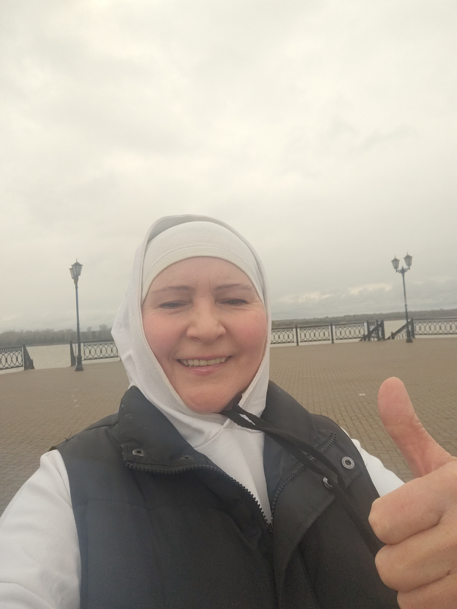 A woman with fair skin and a light-colored head covering is holding the camera and taking a selfie while giving a thumbs-up. She is wearing a white hoodie and a black vest. Behind her is a brick-paved embankment with a decorative metal fence, lamp posts, and a body of water, likely the Kama River. Across the water, trees and the opposite bank are visible. The sky is overcast. The location is likely in the city of Sarapul, Russia, based on the visible architecture and the landscape.