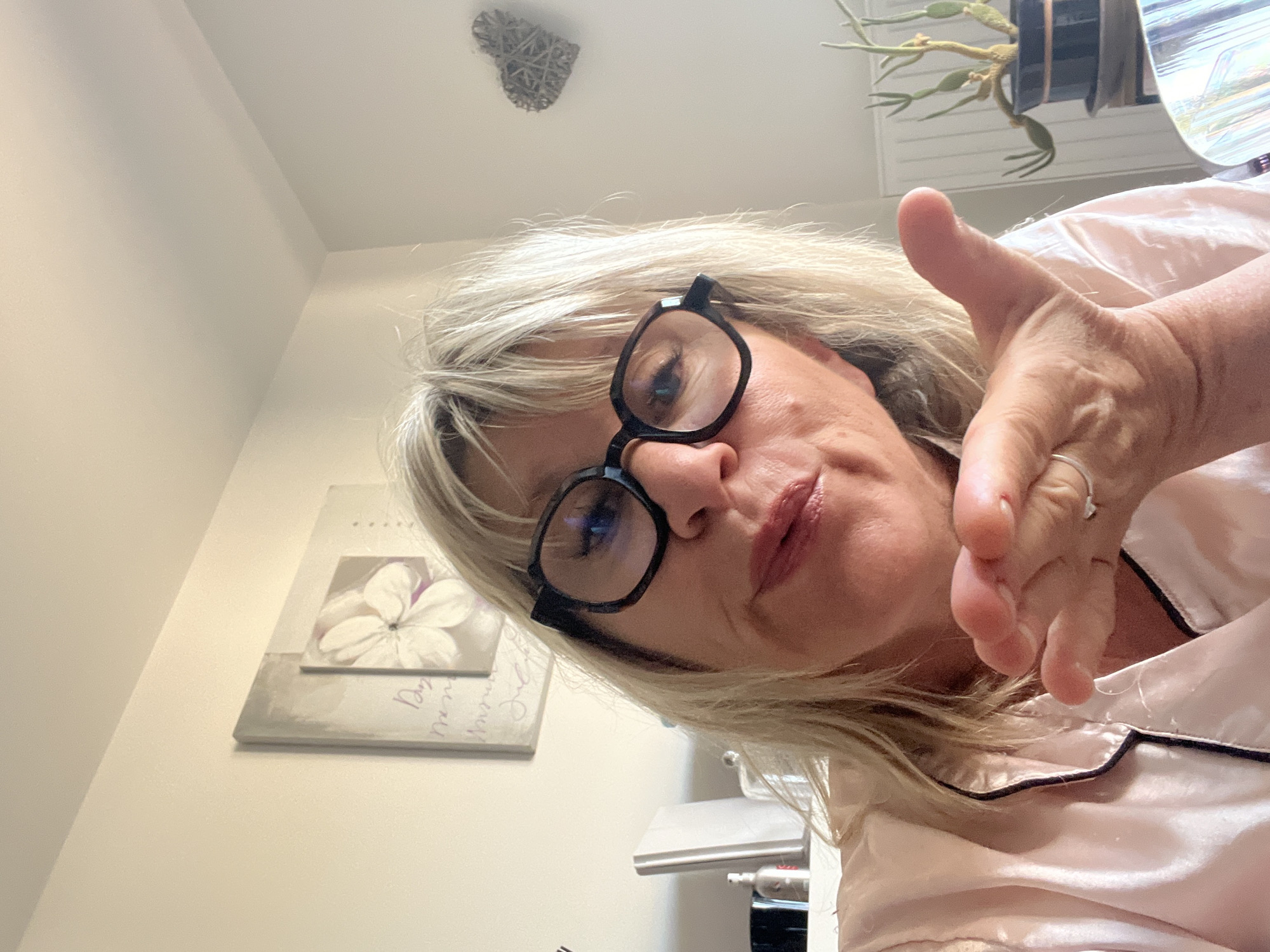 This is a selfie of a woman with blonde hair and glasses, wearing a pink satin pajama top. She is blowing a kiss towards the camera with her right hand extended. She has a ring on her left ring finger. In the background, there is a painting of a flower on the wall, a wicker heart decoration, and a potted plant. The scene appears to be indoors, likely a bedroom. No specific location in Chambœuf, France can be determined from the image.