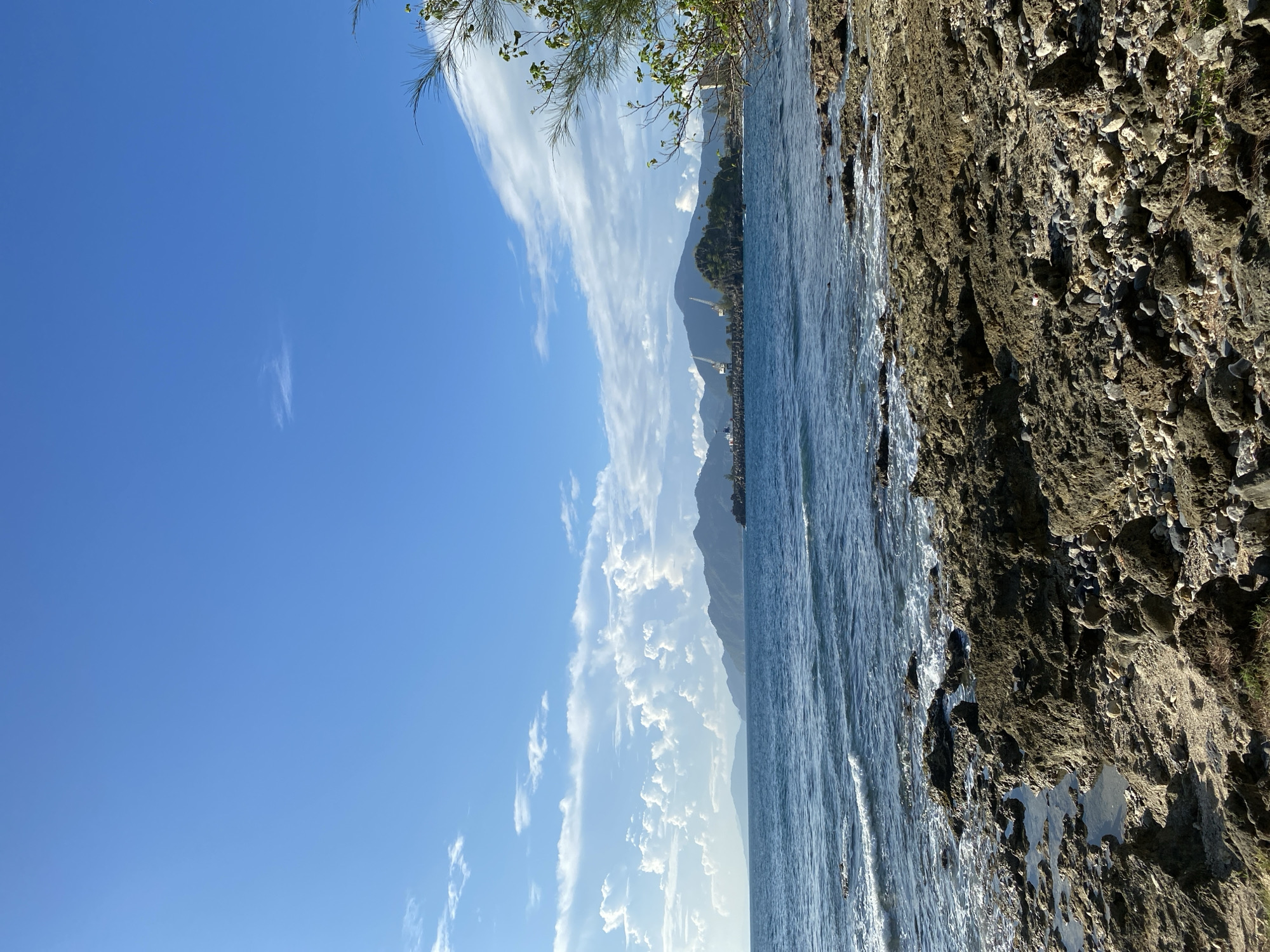 The image depicts a rocky shoreline in Naga Umbang, Indonesia, under a clear blue sky with scattered white clouds. The foreground is dominated by rugged, grey and brown rocks, with some small tide pools visible. The ocean is a deep blue, with gentle waves lapping at the shore. In the mid-ground, a distant coastline with mountains is visible, dotted with some structures that appear to be part of a port, including cranes. A lone tree with green foliage is seen on the right side of the frame, extending from the upper right corner. There are no people visible in the image. The overall scene is one of a natural coastal landscape with hints of industrial activity in the distance.