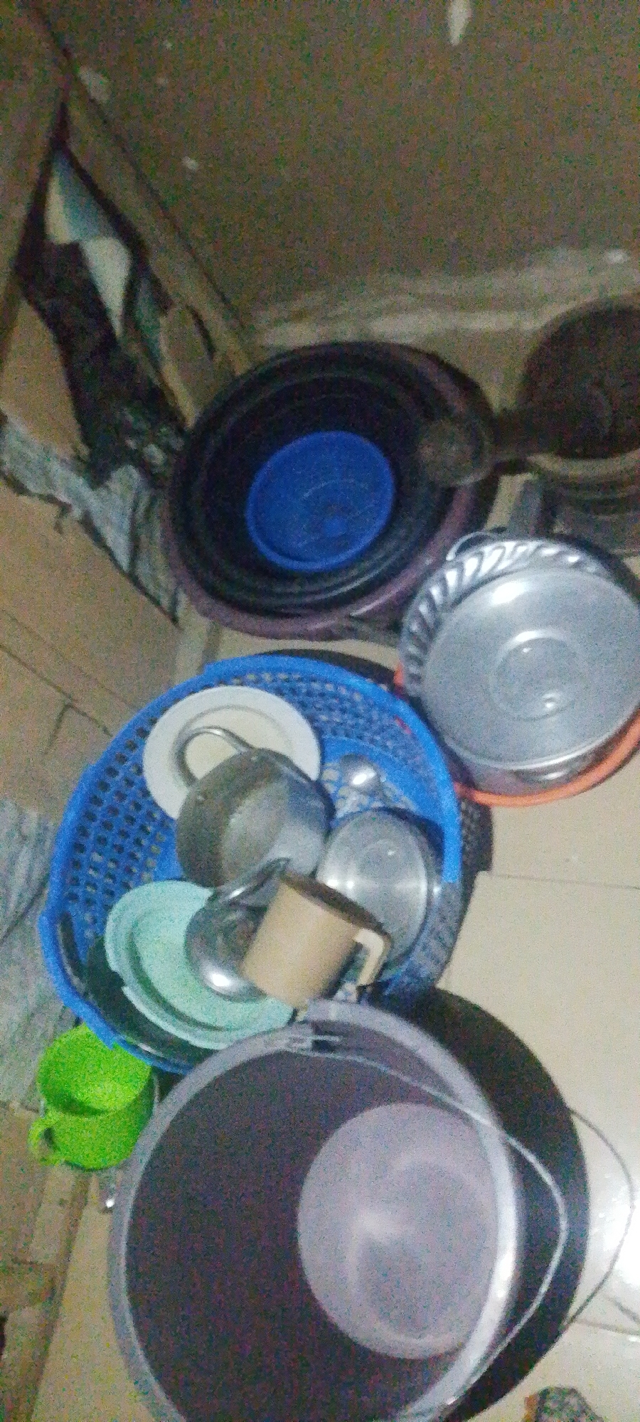 In Jauro Yinu, Nigeria, a collection of dishes and containers is arranged on a floor. A large, dark circular object, possibly a basin or tub, is positioned in the upper portion of the image, with a blue lid or insert visible within it. Below this, a blue plastic basket holds several metal pots and plates. To the left of the basket, a bright green plastic cup is visible. In the foreground, a large, dark circular container, perhaps a bucket, is present. The overall scene appears to be an indoor domestic setting, likely a kitchen or utility area.