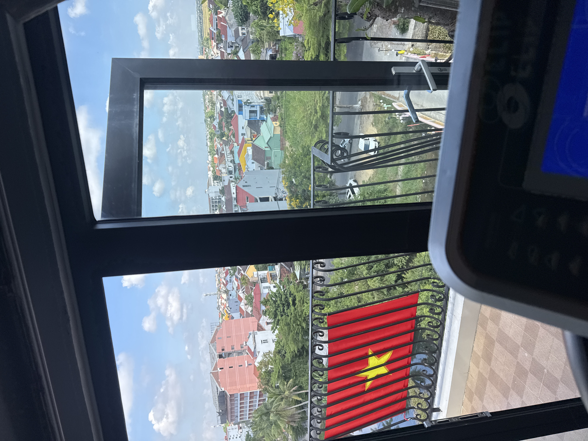 Captured from an elevated indoor viewpoint, the scene features a segment of Hội An Ward, Vietnam, partially framed by the structure of a window or doorway. A prominent Vietnamese flag is displayed on a balcony railing. The cityscape unfolds with a mix of residential and commercial buildings, interspersed with verdant trees. In the immediate foreground, a piece of equipment, possibly a treadmill, occupies the right side of the frame. The sky is blue with scattered clouds.