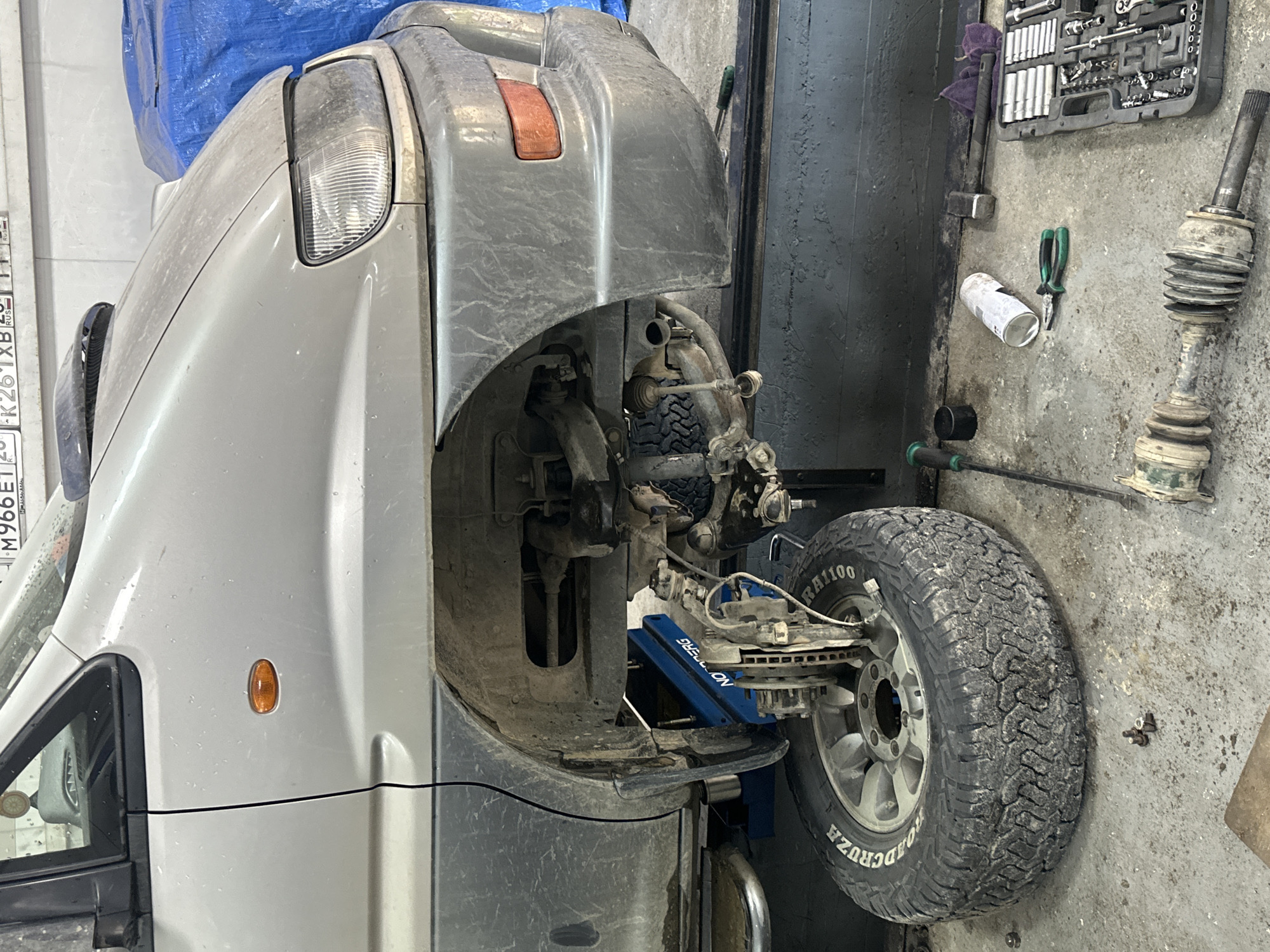 The image depicts a vehicle, likely a silver minivan, undergoing repair in what appears to be a garage or workshop in Blagoveshchensk, Russia. The front passenger side of the vehicle is raised on a blue hydraulic lift, and the wheel has been removed, revealing the brake assembly and suspension components. Various tools are scattered on the concrete floor, including a socket wrench set in a grey case, a can of spray lubricant, pliers, a hammer, and what appears to be a removed driveshaft. The environment is dusty, suggesting an active workspace. The license plate visible on the car reads "M966ET" with Russian characters below. There are no people visible in the image.