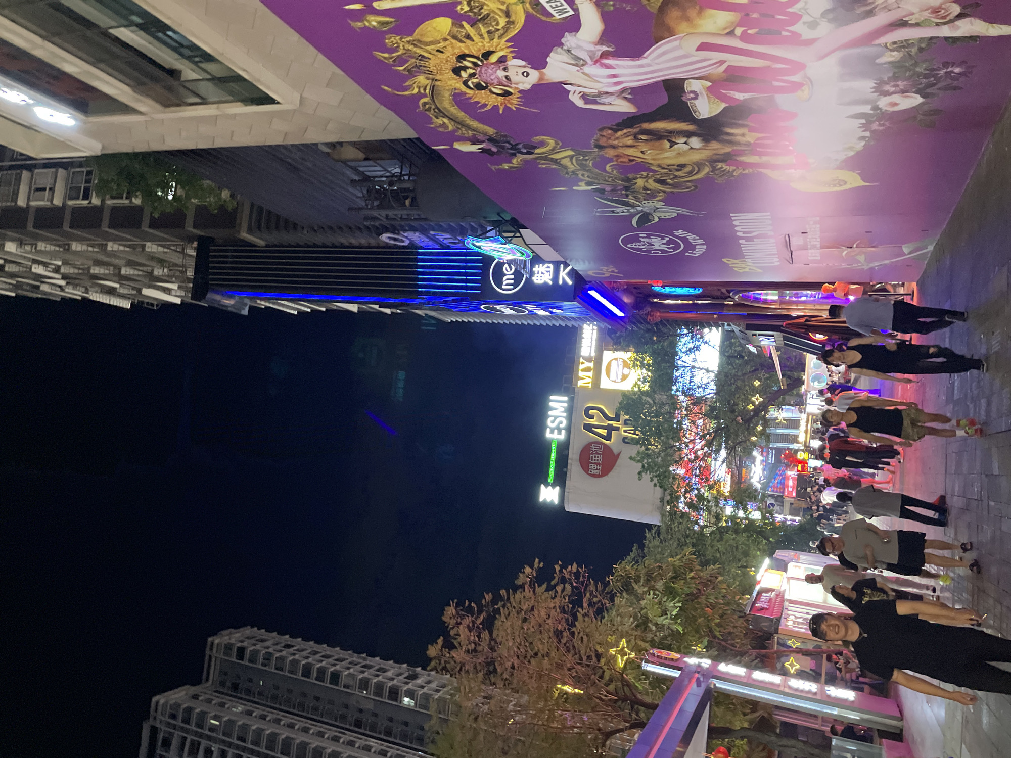 The image depicts a street scene at night in the Liangjiang New Area of Chongqing, China. The street is lined with tall buildings and illuminated by neon signs and streetlights. Several people are walking on the sidewalk, some in groups and others individually. One group of young men is walking towards the camera, while other pedestrians are seen further down the street. A large, colorful mural is painted on a wall to the right of the street, featuring a woman in a flamboyant headdress and a lion. Signs indicate that a "BAR COMING SOON" in this area. The overall atmosphere is vibrant and busy, suggesting a lively urban environment.
