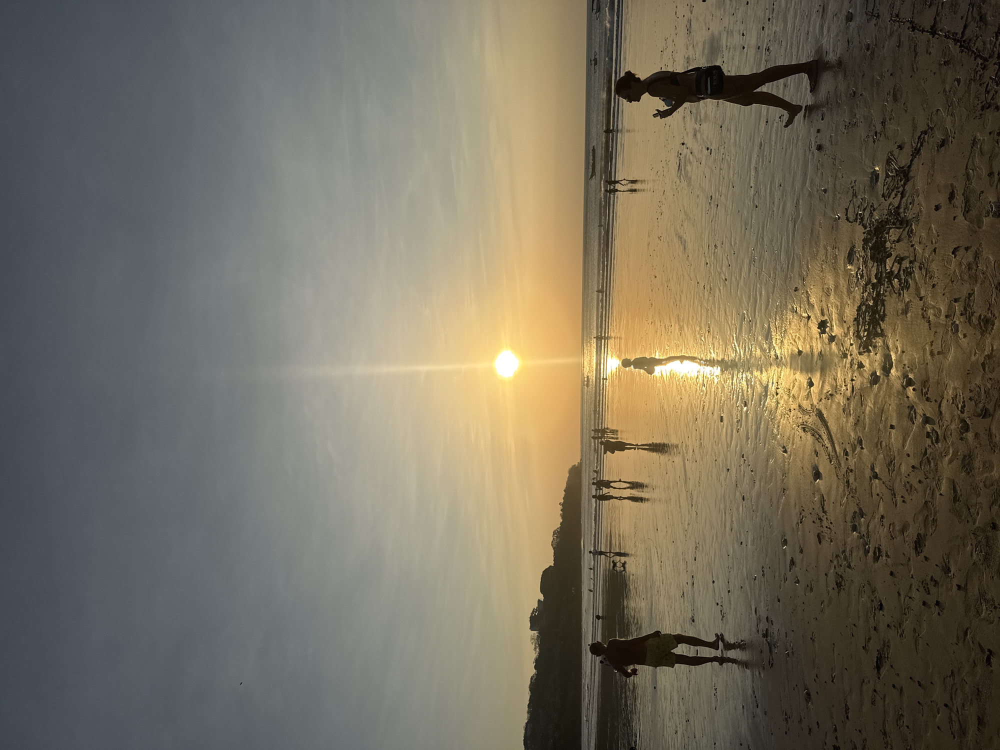A wide-angle vertical shot captures a serene sunset over Kuta Selatan, Indonesia's Kuta beach. The sun, a bright disc, hangs low in the sky, casting a golden reflection across the wet sand. The sky is a gradient of soft blues and yellows, with a few wispy clouds. Silhouetted figures dot the beach, some walking along the shoreline, others gathered in small groups. The ocean is calm, with gentle waves lapping at the shore. A few boats are visible in the distance. The sand is speckled with footprints and small shells, indicating recent activity. The overall mood is peaceful and tranquil, typical of a sunset on a popular tourist beach.