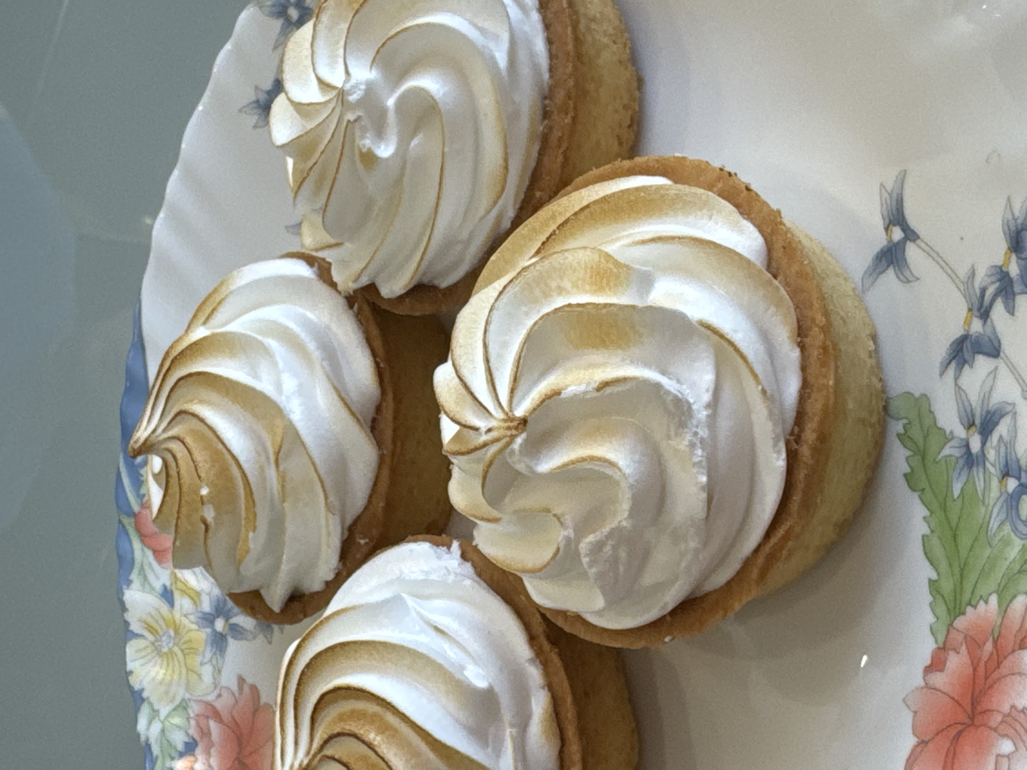 The image shows four lemon meringue tarts arranged on a decorative plate with floral patterns. The tarts have a golden-brown crust and are topped with swirls of white meringue that is slightly toasted at the tips. The floral pattern on the plate includes blue and red flowers. The image does not provide any information about the location in Saint Petersburg, Russia. There are no people visible in the image.