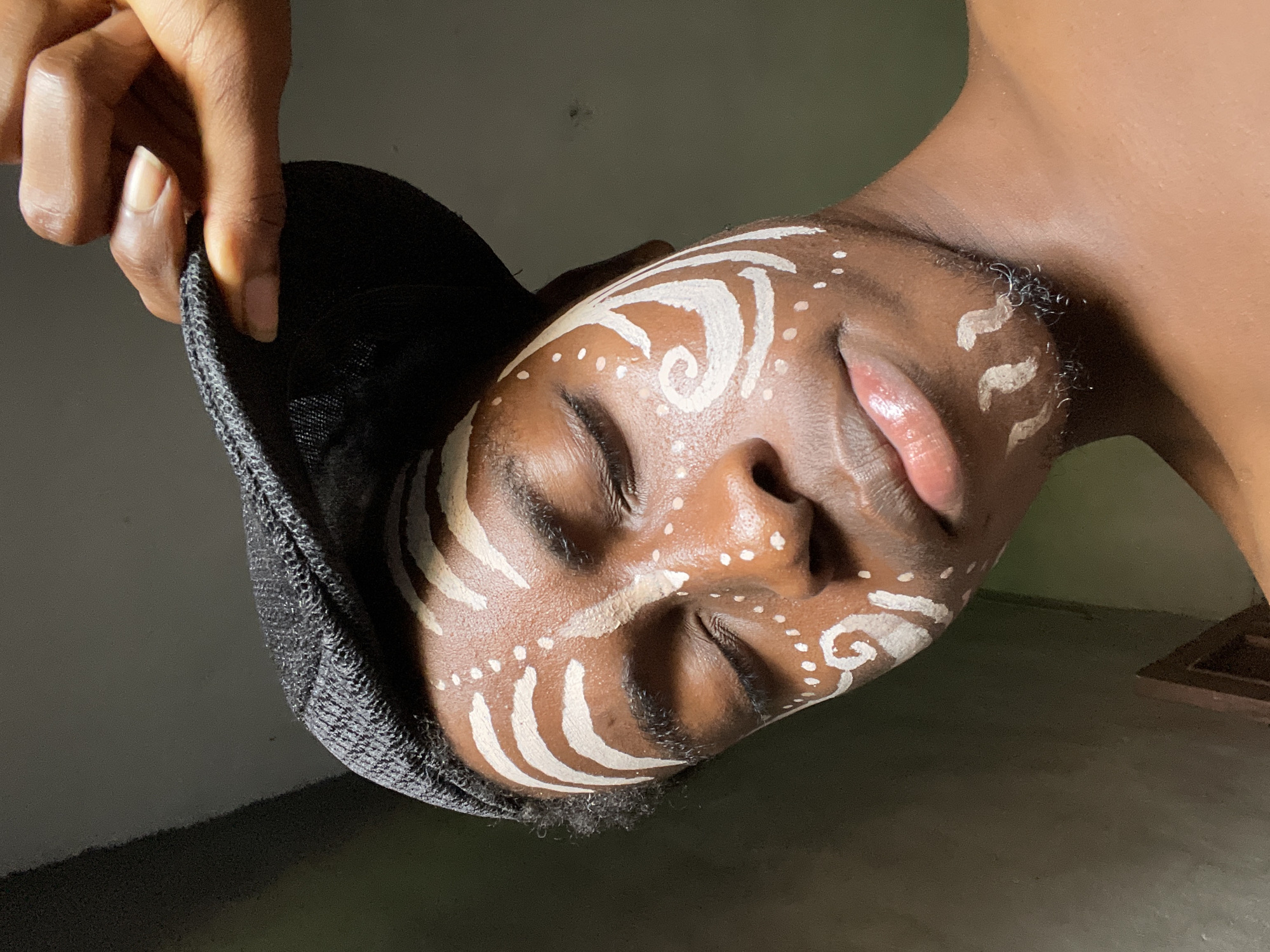 The image captures a person with dark skin, their head tilted significantly to the left, eyes closed, conveying a serene or meditative expression. Their face is adorned with striking white patterns, including spirals, swirls, and numerous small dots, applied across the forehead, cheeks, nose, and chin. A hand with dark skin and neat nails gently grasps the brim of a dark, textured cap or hat, holding it near the person's head. The person's bare shoulder and neck are visible, suggesting an unclad upper torso. Short, dark facial hair is visible along the jawline and chin. The background is simple and dark, appearing to be an indoor wall or a dimly lit, plain surface, with a faint suggestion of a floor or a lower surface towards the bottom right. The lighting is soft, highlighting the contours of the face and the details of the face paint. No text is visible in the image. The scene is set in Nkpor, Nigeria.