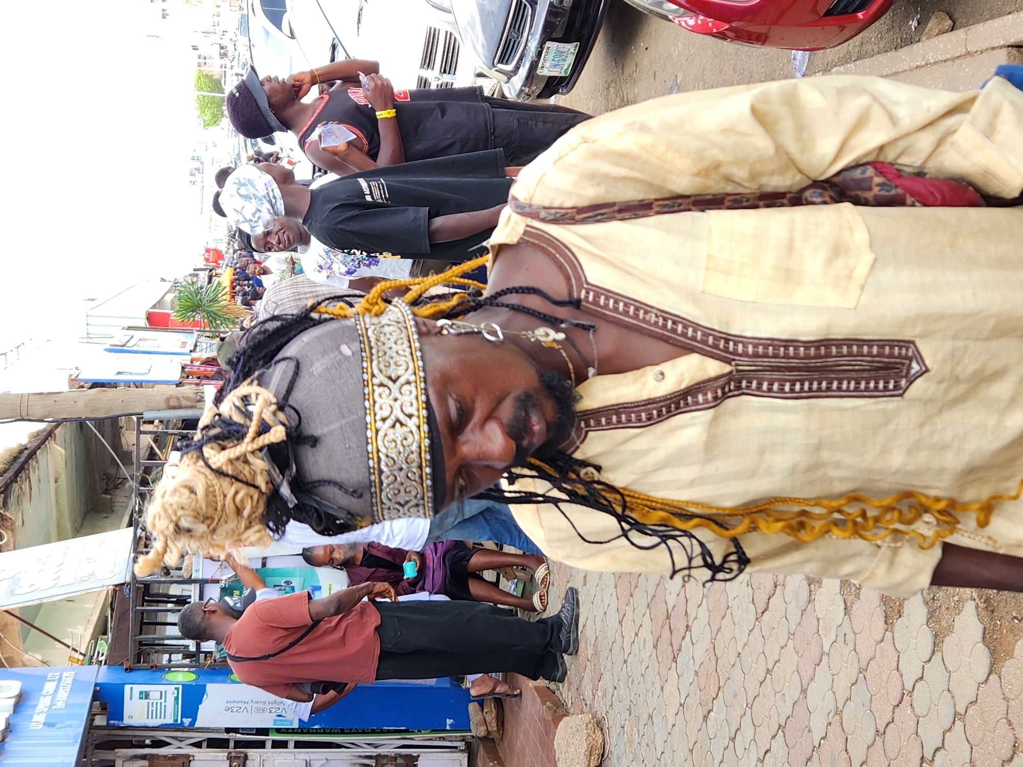 Here's a detailed description of the image:
**Scene:** An outdoor scene on a paved area resembling a sidewalk or public space. The image appears to be taken in an urban setting, with buildings and vehicles visible in the background.
**People:**
* **Foreground:** A man wearing a light-colored, traditional-style shirt with brown trim, a head covering adorned with intricate patterns and decorative braids or cords (black and yellow), and a necklace. His eyes appear closed, and he has a beard and mustache.
* **Background:** Several other individuals are visible in the background, including:
* A man wearing a red shirt and black pants. He's holding a piece of paper.
* Several other people, dressed in casual clothing, appear to be standing or walking.
**Objects:**
* Several Cars are parked, the one in front of the image has license plate number: UNG6399DJ
* There's a sign or display board on the left side of the image, it contains the phrases "THREE DAYS WARRANTY" and "Every Moment", and the word "VIVO".
* There are rocks around the pavement.
**Actions and Interactions:**
* It's difficult to determine specific actions, but the people appear to be in a public gathering or event. The man in the foreground is in the center of attention, which may indicate he is at the parade.
**Environment:**
* The setting is urban with buildings and vehicles.
* The pavement is made of interlocking stones or tiles.
**Location:** The specific city or location is not identifiable from the image, though it's indicated as Unknown, Unknown.