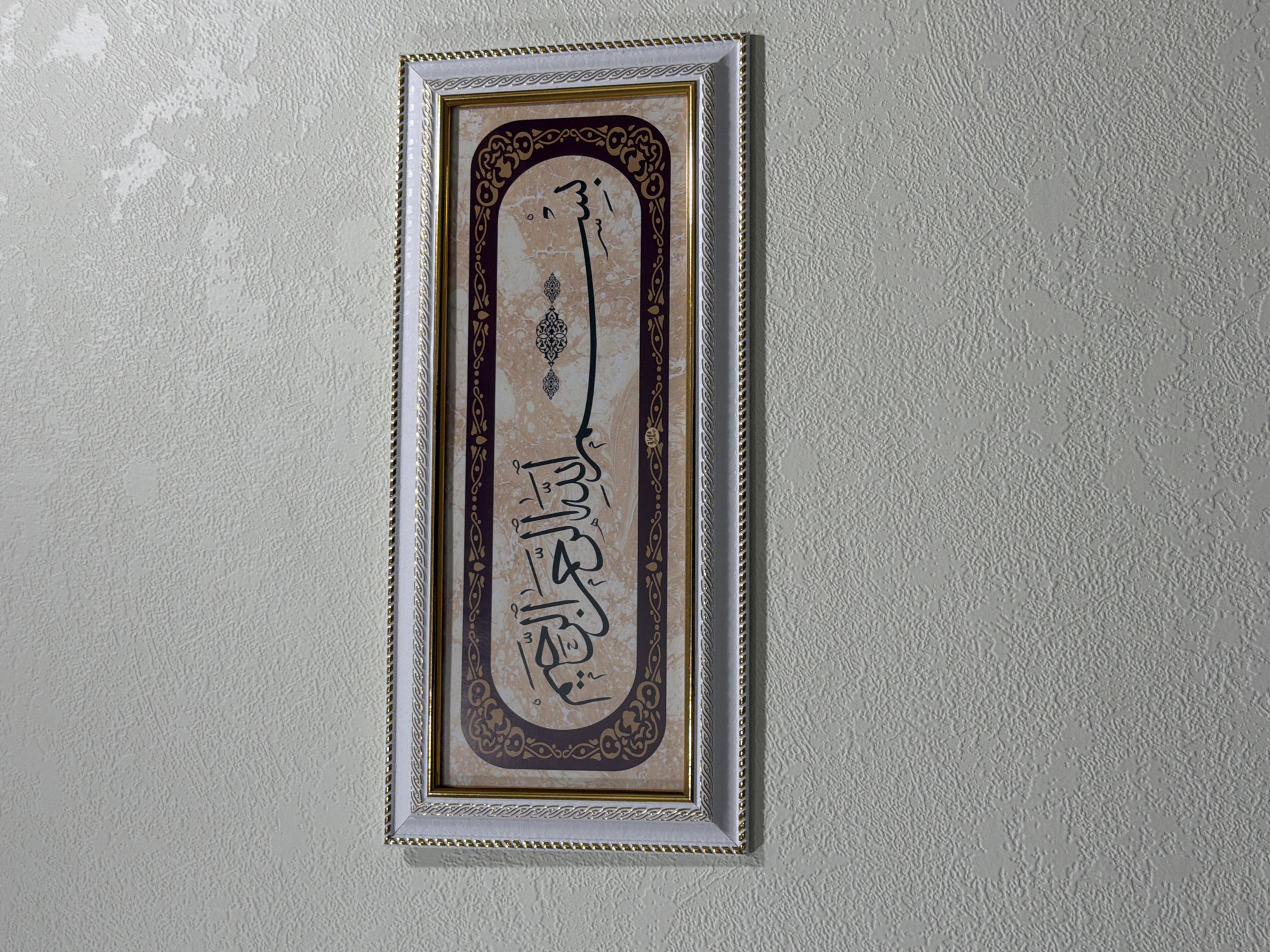 A framed Islamic calligraphy artwork hangs on a textured, light-colored wall. The artwork features Arabic script in black ink on a marbled background, surrounded by a decorative border. The frame is white with a gold trim and a braided rope detail. The image is taken from a slightly low angle, focusing on the artwork and the wall it is mounted on. There are no people or animals visible in the image.