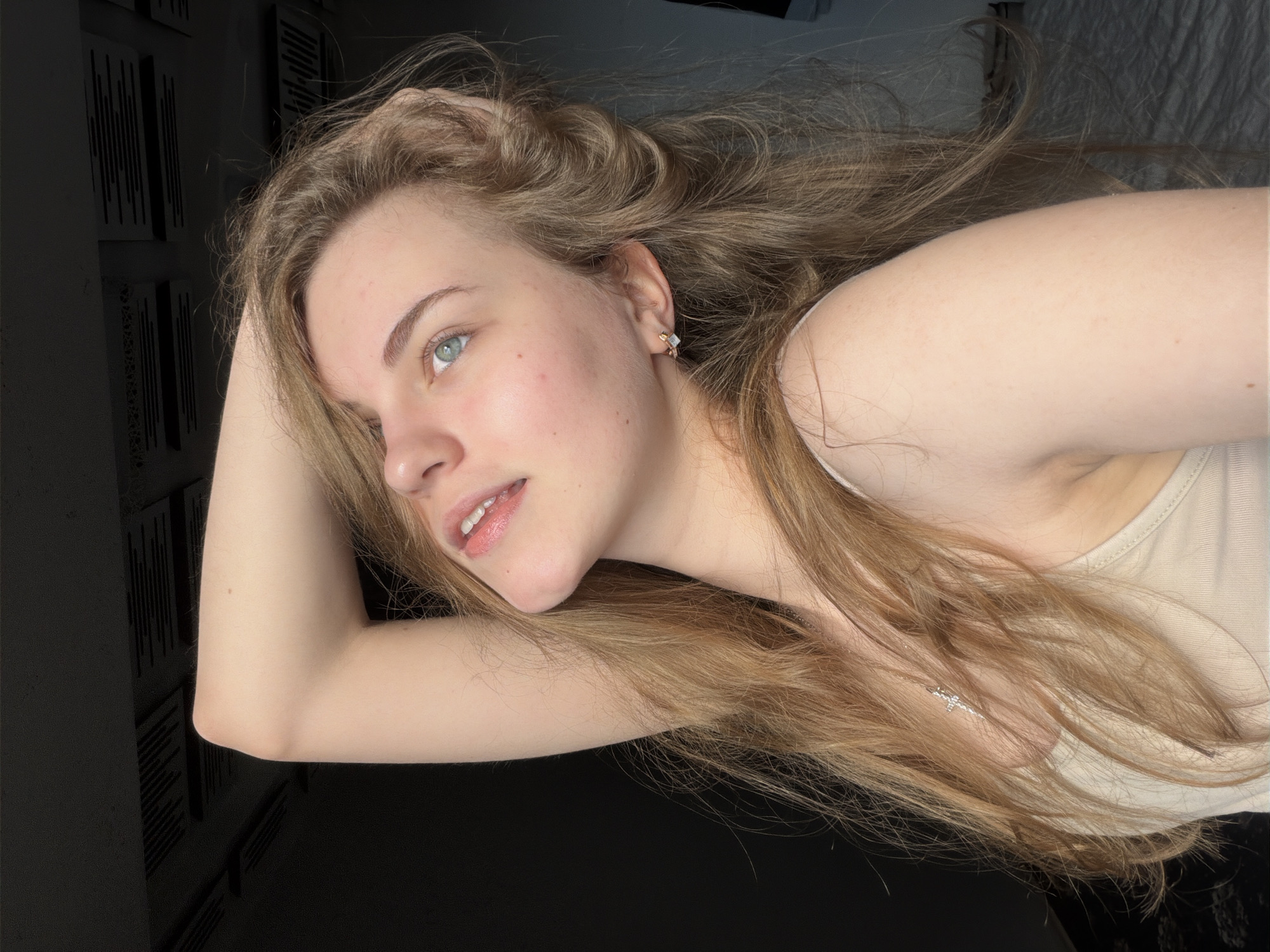 The image is a close-up, low-angle, self-portrait of a young woman with blonde hair and blue eyes. She is wearing a beige tank top and a silver cross necklace. Her right arm is raised behind her head, and her left arm is not visible. She is looking up and to her left, with her mouth slightly open. The background is dark, with some patterned ceiling tiles visible at the top. There is no discernible location in the city of Dnipro, Ukraine.