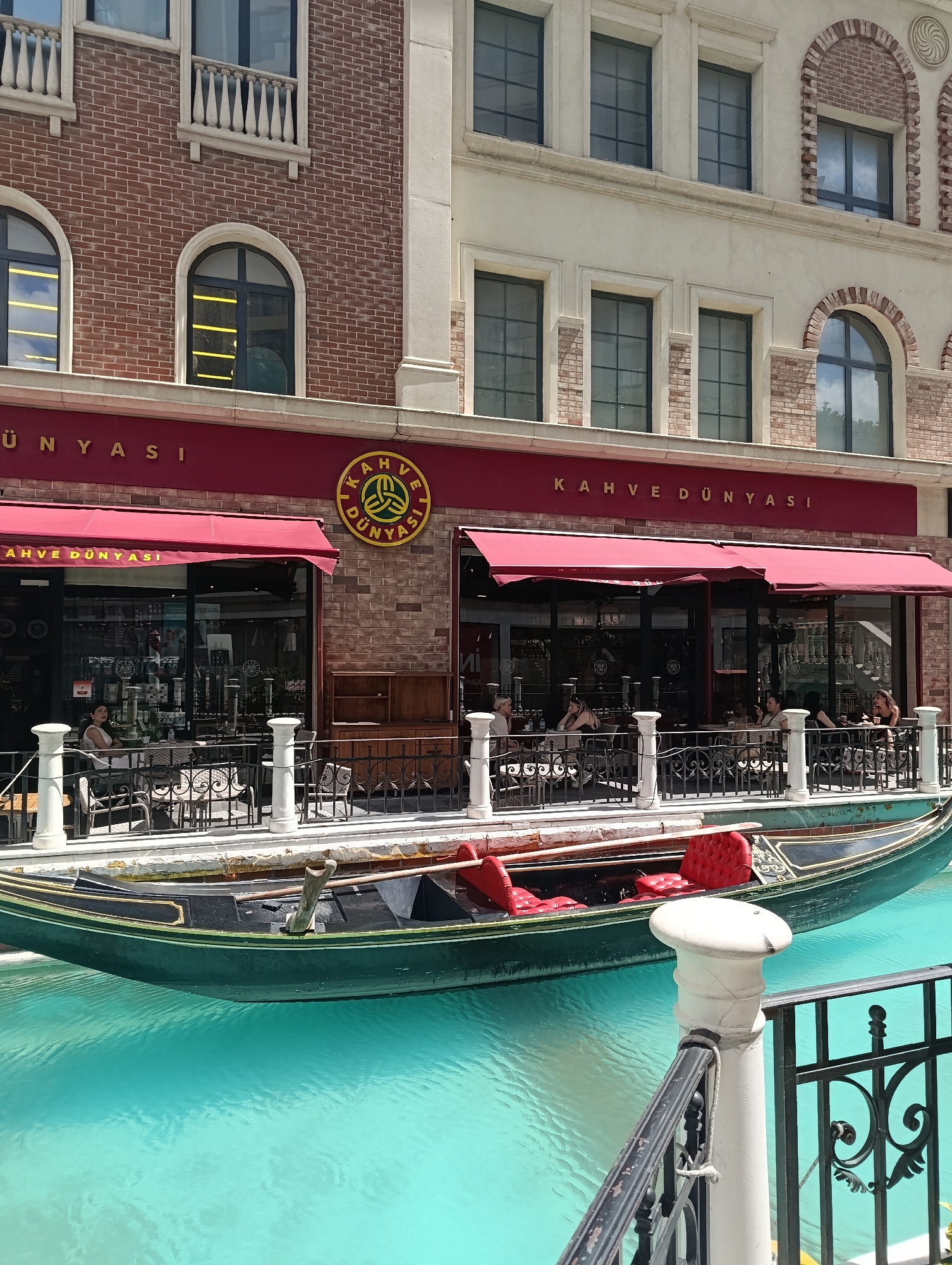 The image captures a picturesque outdoor seating area of a "Kahve Dünyasi" cafe, situated in the Venezia Mega Outlet mall in Gaziosmanpaşa, Istanbul, Turkey. The cafe is characterized by its red awnings and large glass windows, offering a view of a turquoise canal. A traditional Venetian gondola, painted dark green with gold trim and red velvet seats, floats in the canal, adding a unique touch to the setting. Several patrons are seated at tables on the outdoor patio, enjoying their drinks and the ambiance. The architecture of the surrounding buildings, with their brick facades and arched windows, evokes an Italian style, mirroring the Venice theme of the mall. White balustrades and decorative railings line the canal, creating a safe and elegant promenade. The scene is bathed in natural daylight, suggesting it was taken during the daytime.