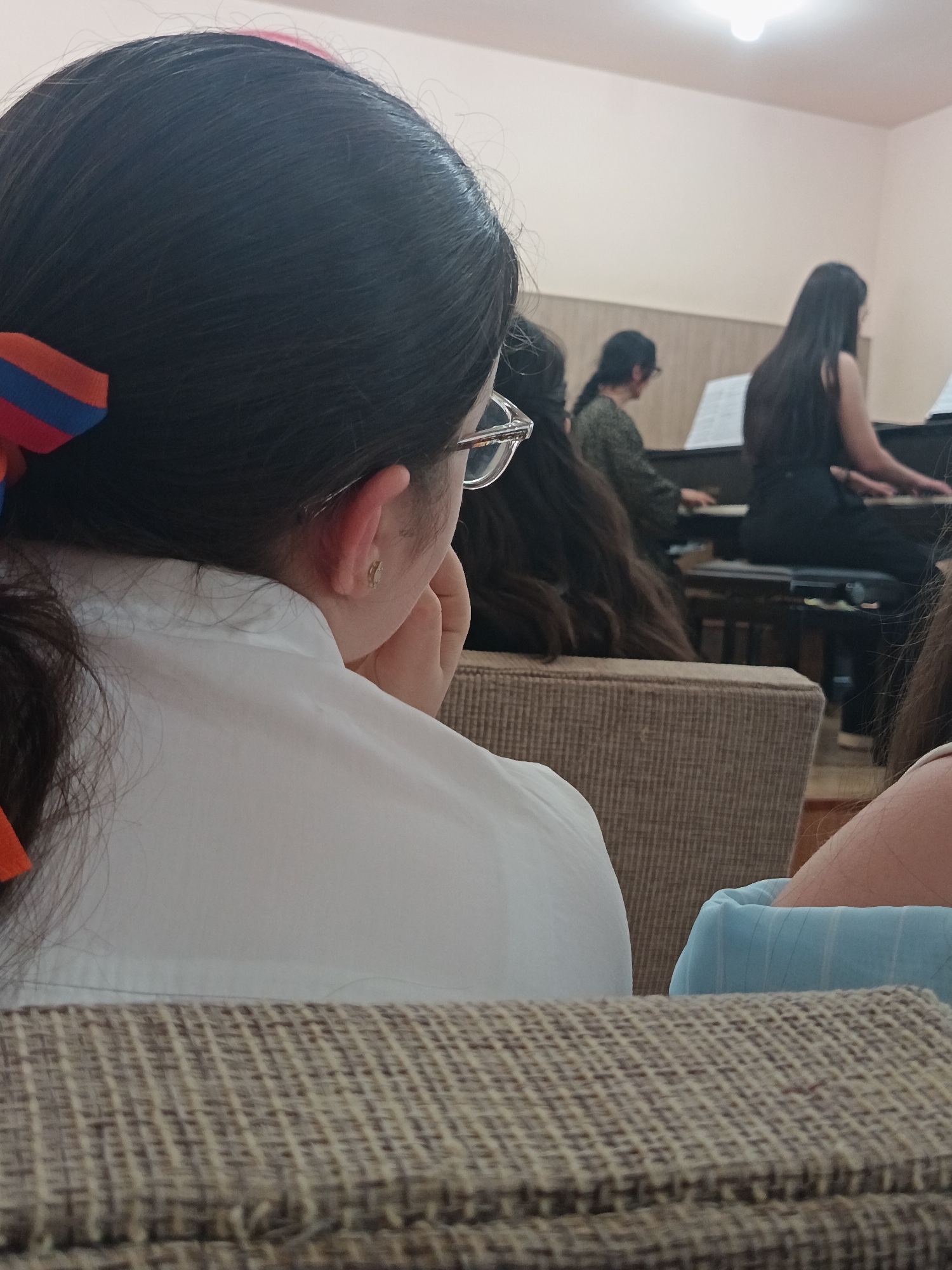 In an indoor setting, likely a music classroom or performance space, the image captures several individuals. In the foreground, a young person with dark hair, wearing glasses and a white shirt, is seen from behind, resting their chin on their hand. They have an Armenian flag ribbon in their hair. Behind them, two other individuals are seated at a piano, with sheet music visible. The environment appears to be a dimly lit room with light-colored walls and furniture. The overall impression is that of a musical rehearsal or lesson.