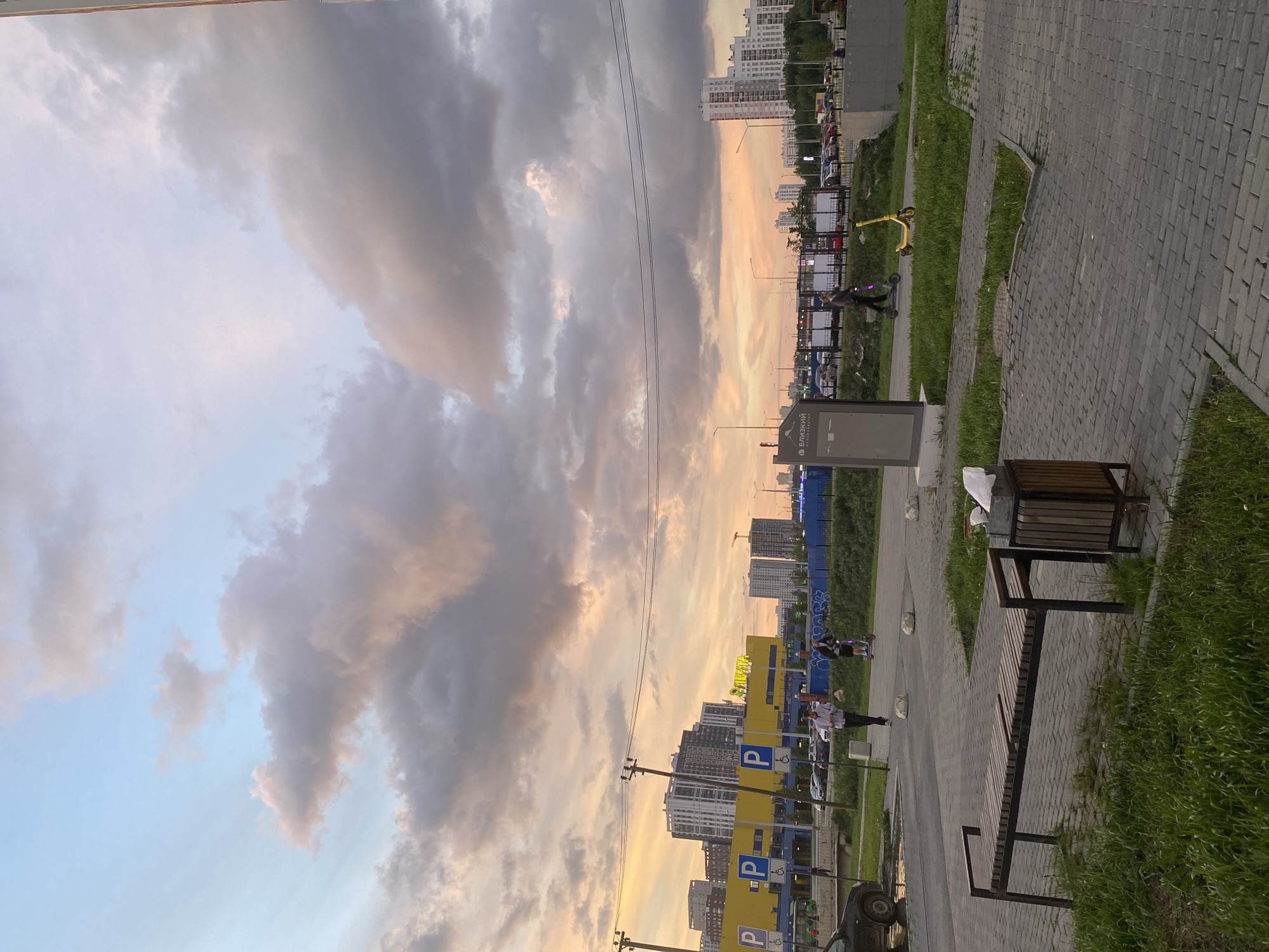 This image captures an urban or suburban landscape, likely in Yekaterinburg, Russia, during the late afternoon or early evening, characterized by the warm, golden light of a low sun. The sky is a prominent feature, filled with large, dramatic clouds displaying a mix of soft pink, orange, and gray tones, contrasting with patches of light blue sky. Power lines are visible stretching across the upper portion of the frame.
The midground is dominated by a series of modern, multi-story residential buildings, mostly white or light-colored, suggesting a contemporary development. Some construction cranes hint at ongoing development. A street or parking area runs alongside these buildings, with numerous parked cars visible. Bright yellow signs with a blue "P" symbol, indicating parking, are frequently spaced, some also displaying a wheelchair accessibility symbol. One sign prominently features the Russian text "БЛИЗКИЙ КВАРТИРЫ" (Blizkiy Kvartiry), which translates to "Near Apartments," confirming the residential context.
In the foreground, a wide paved pedestrian path, constructed from gray interlocking bricks, meanders through the scene. It is bordered by vibrant green grass on both sides. On the right, a long wooden bench with a dark metal frame is placed beside a matching trash can. A gray information or advertising stand is also visible nearby. Several individuals are present: one person is riding a dark electric scooter on the grassy area, while a bright yellow scooter rests unattended nearby. Other people are seen walking or standing further down the path, near a blue fence. The overall scene depicts a calm, everyday setting within a developing urban environment.