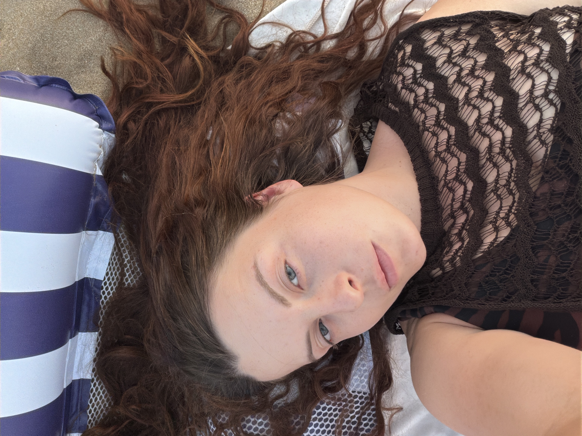 A woman with long, wavy, auburn hair lies on her back on a beach, with her head resting on a striped inflatable pillow. She wears a dark brown, crocheted top over a patterned garment. Her eyes are open and looking at the camera. The environment suggests a beach setting in Manavgat, Turkey, with sand visible in the background and a mesh-like surface beneath her.