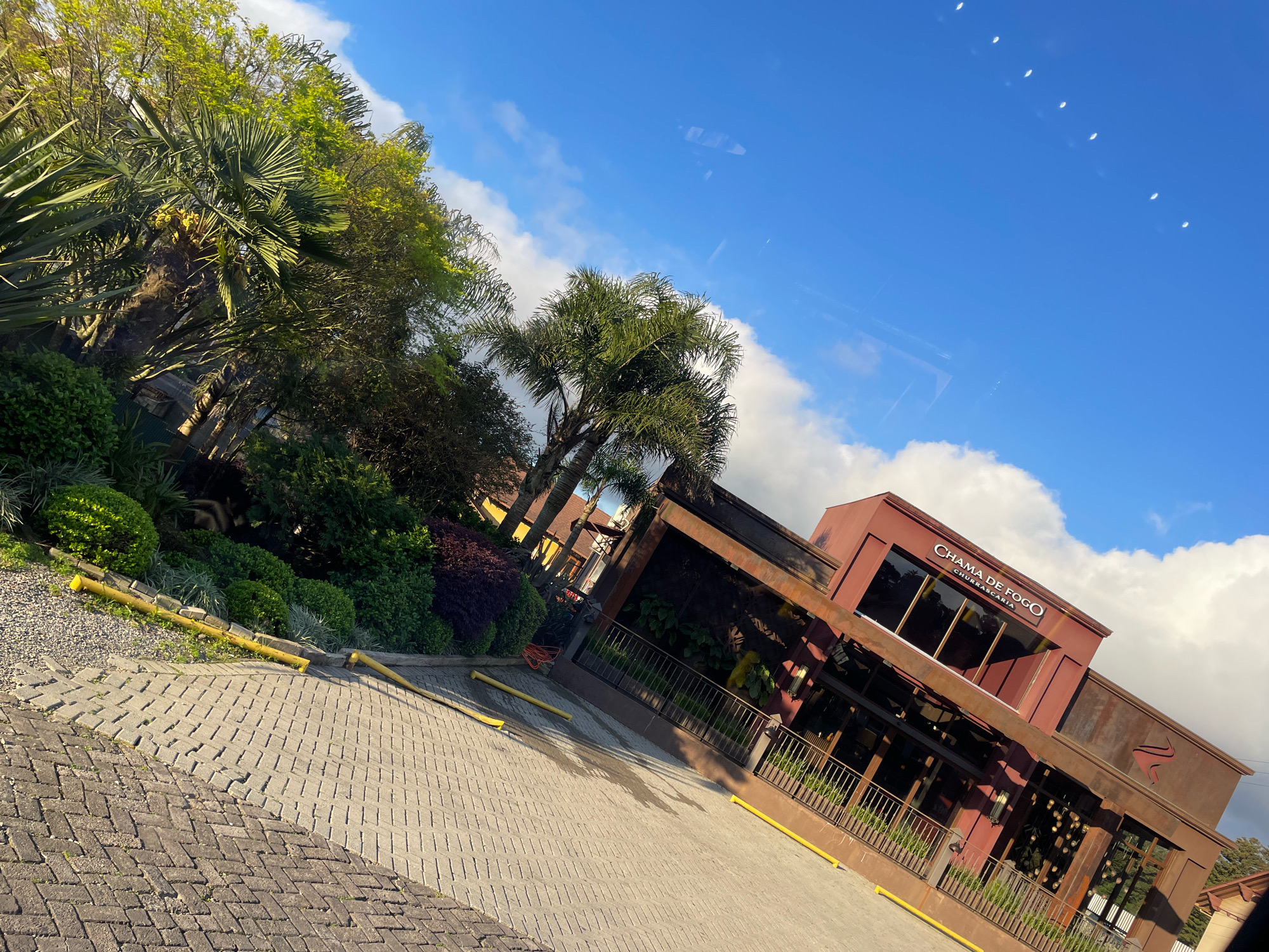 The image shows the exterior of a restaurant named "CHAMA DE FOGO CHURRASCARIA" located in the city of Gramado, Brazil. The building has a rustic, modern design with dark brown, weathered metal accents and a deep red-colored facade. Large windows are visible, and the restaurant's name is prominently displayed above the entrance. In front of the building, there is a paved area with yellow bollards marking parking spaces. Lush green landscaping, including palm trees and various shrubs, surrounds the building. The sky above is blue with scattered white clouds, and the sunlight suggests it is daytime. The photograph is taken from a slightly elevated angle, giving a view of the building, its surroundings, and a portion of the sky. The ground in the foreground is paved with interlocking bricks arranged in a herringbone pattern. There are no people visible in the image.