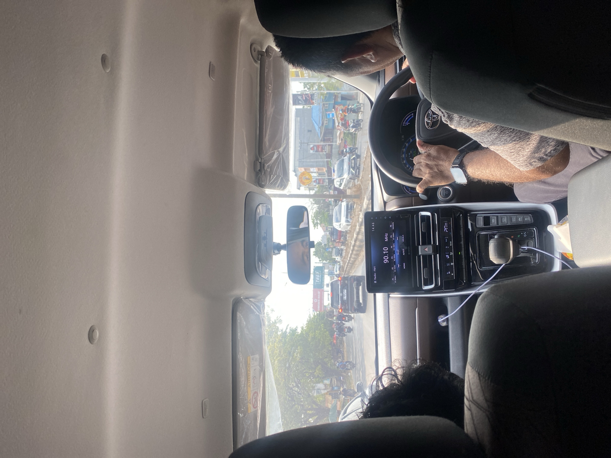A POV shot from the interior of a car shows a driver in the front passenger seat of a car in the city of Gambut Barat, Indonesia. The driver, a man with short hair and wearing a gray patterned shirt, holds the steering wheel with his right hand. The man is wearing a watch on his left wrist. The car's infotainment system displays the radio station "90.10 MHz" and the time "14:46." Outside the car, the street is busy with traffic, including motorcycles and cars. Buildings and trees line the roadside. The sun is shining, casting shadows.