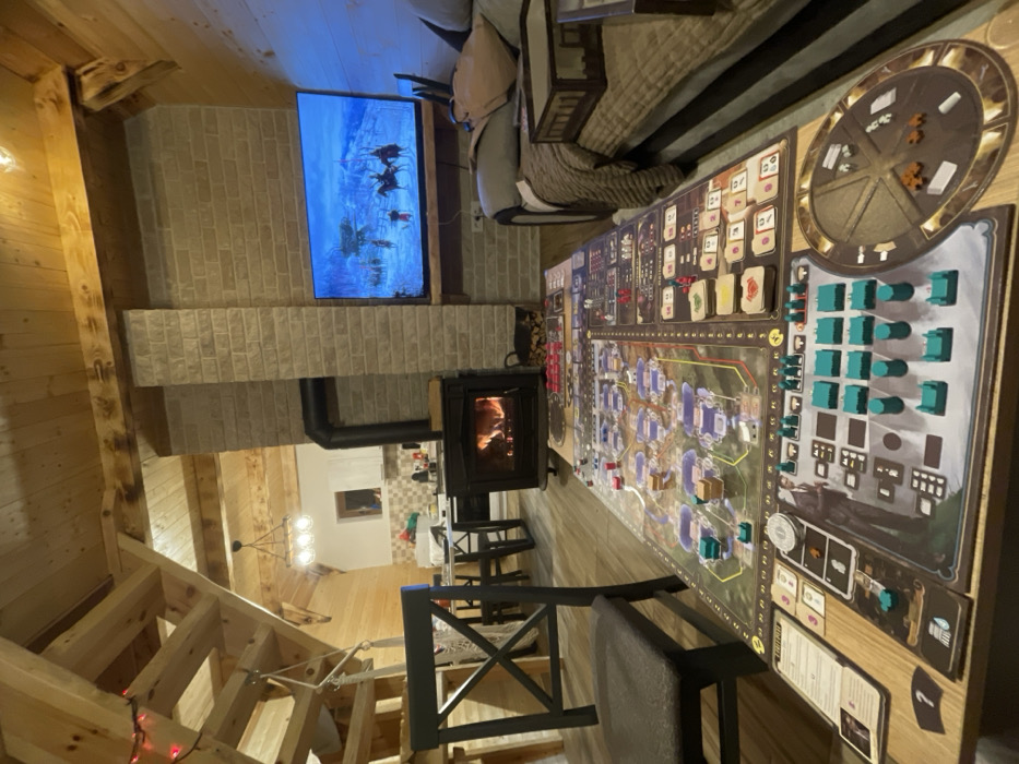 This image displays an interior view of a living space within a dwelling in Palež, Montenegro. The central feature is a large, multi-component board game, identified as "Barrage," fully arranged on a wooden coffee table. The game setup includes numerous teal, red, white, and brown tokens, cards, and miniature structures spread across the board's various sections. To the immediate left, a dark-stained wooden chair with a cushion is partially visible. To the right, a sofa draped with brown blankets and pillows holds a small, rectangular wooden tray. Directly behind the coffee table, a black wood-burning stove contains active flames and is positioned against a light-toned brick wall. A stack of cut firewood rests on the floor beside the stove. Above the stove, a flat-panel television is mounted, displaying a winter landscape with figures on horseback. The room features exposed wooden ceiling beams and paneling, with a wooden staircase ascending on the left, adorned with a string of red lights and a decorative hanging element. In the background, a kitchen area with white cabinetry, a counter, and a dining table with chairs is visible, illuminated by a multi-bulb chandelier. No individuals are present in the scene.