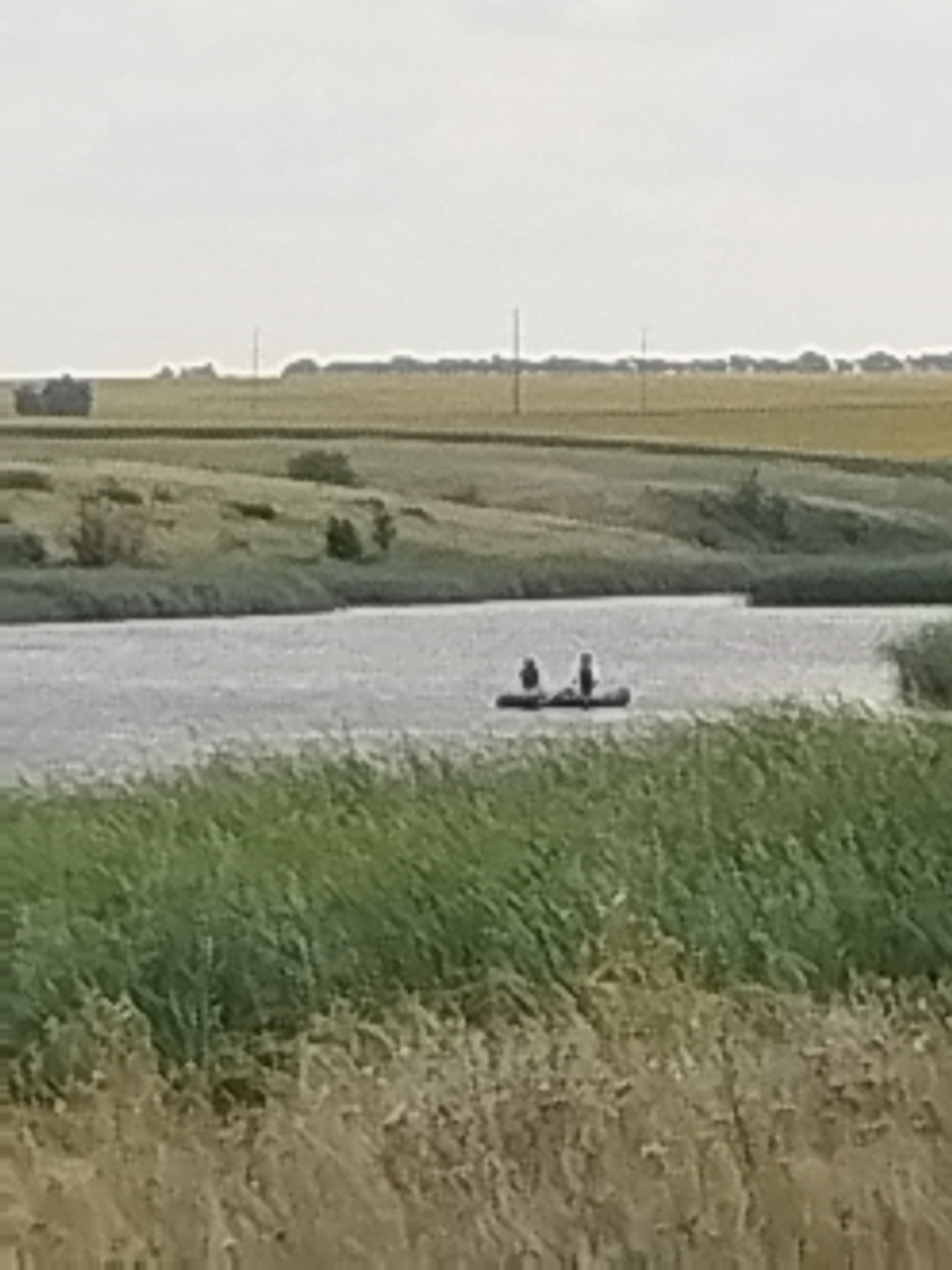 Two individuals are seated in a narrow boat on a body of water, viewed from across a grassy bank. The boat is dark and elongated, with the individuals appearing as dark figures within it. The water is choppy and reflects the overcast sky. In the background, rolling hills of green and yellow vegetation stretch towards a hazy horizon. A few scattered structures, possibly farm buildings or silos, are visible on the distant hills. The foreground is dominated by tall, dry grass, obscuring the lower portion of the scene. The overall impression is of a rural, possibly agricultural landscape under a muted sky. The location is the city of Parfenovka, Russia.