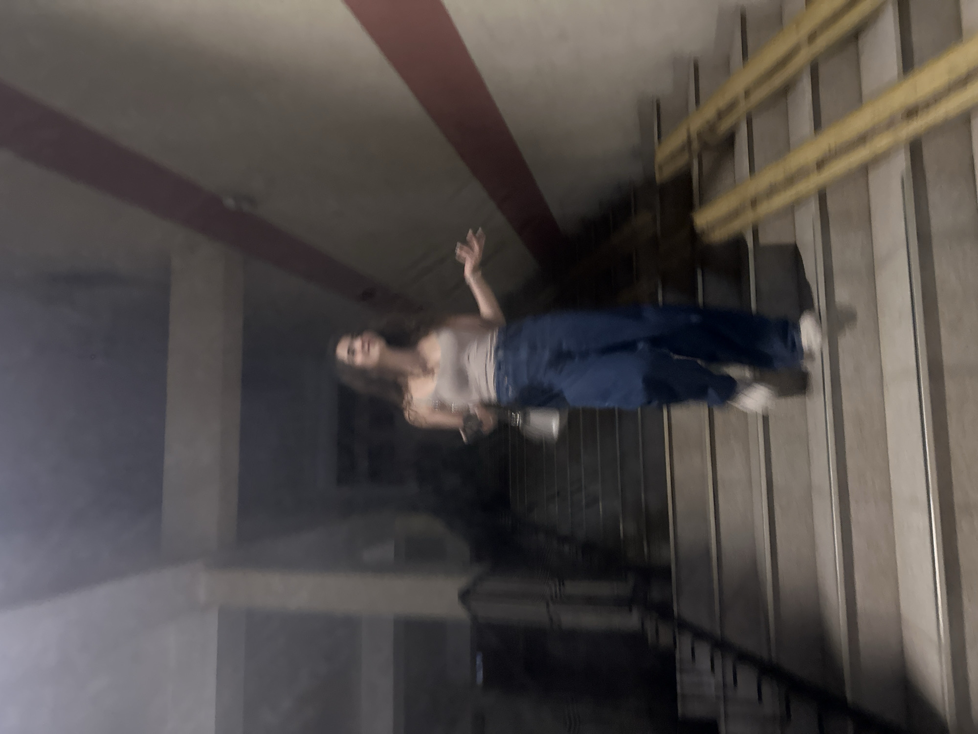 The image depicts a young woman on a staircase, likely indoors, with a motion blur indicating movement. She is wearing a light-colored tank top and baggy blue jeans, and holding a small, light-colored purse. Her expression appears to be one of surprise or curiosity, with her mouth slightly open and her hands extended. The staircase is made of concrete with metal edging. There are two wooden ramps running down the side of the stairs. The walls are painted a neutral color with a prominent red stripe. The environment suggests a somewhat utilitarian or public space, such as a parking garage or an institutional building. There is no specific indication of the city of Shumen, Bulgaria, within the image itself.