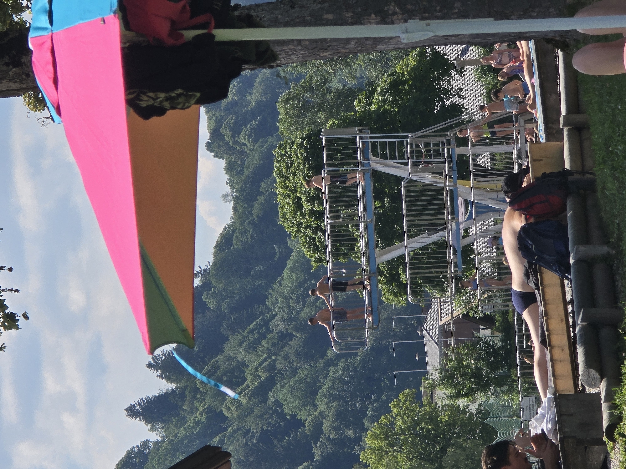In Garmisch-Partenkirchen, Germany, a bright, multi-colored umbrella shades a sun-drenched outdoor swimming pool area. In the foreground, a person lies on a towel, their back to the camera, with a backpack beside them. A man is seated in the left foreground, looking down at a towel.
In the midground, a diving platform with multiple levels stands prominently. Several young boys are visible on the platform; two are standing on the lowest level, facing away, while another is on a higher level. A few other people, including a woman in a bikini and children, are visible in the background near the pool.
The background is dominated by a lush, tree-covered hillside, suggesting the natural beauty of the Bavarian Alps surrounding Garmisch-Partenkirchen. The sky is a clear blue with scattered white clouds, indicating a pleasant, sunny day. A blue and white ribbon or streamer is attached to a pole or structure in the upper left of the frame, fluttering in the breeze. The overall scene depicts a recreational activity at a public swimming facility on a warm day.