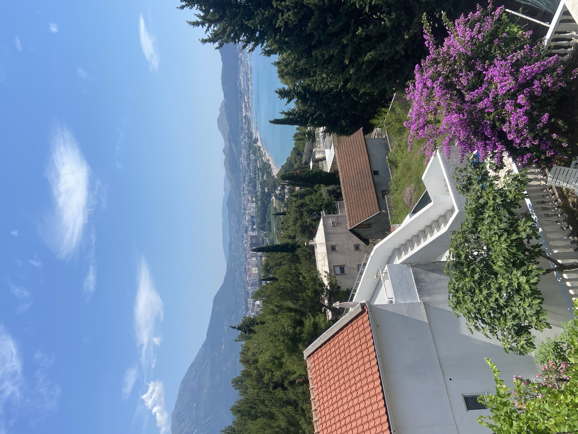 The photograph captures a scenic view from a hillside in Šušanj, Montenegro, overlooking the Adriatic Sea. In the foreground, a white building with a terracotta roof is visible, along with a white staircase leading up to an upper level. Lush green trees and vibrant purple bougainvillea flowers are prominent, adding to the natural beauty of the scene. Beyond the immediate foreground, several other buildings with tiled roofs can be seen, interspersed with more evergreen trees. The middle ground reveals a coastal town with a sandy beach bordering a clear blue sea, dotted with boats in a harbor. In the distance, a range of mountains forms the backdrop, with a hazy sky and scattered white clouds overhead. The overall impression is one of a serene and picturesque coastal landscape on a sunny day.