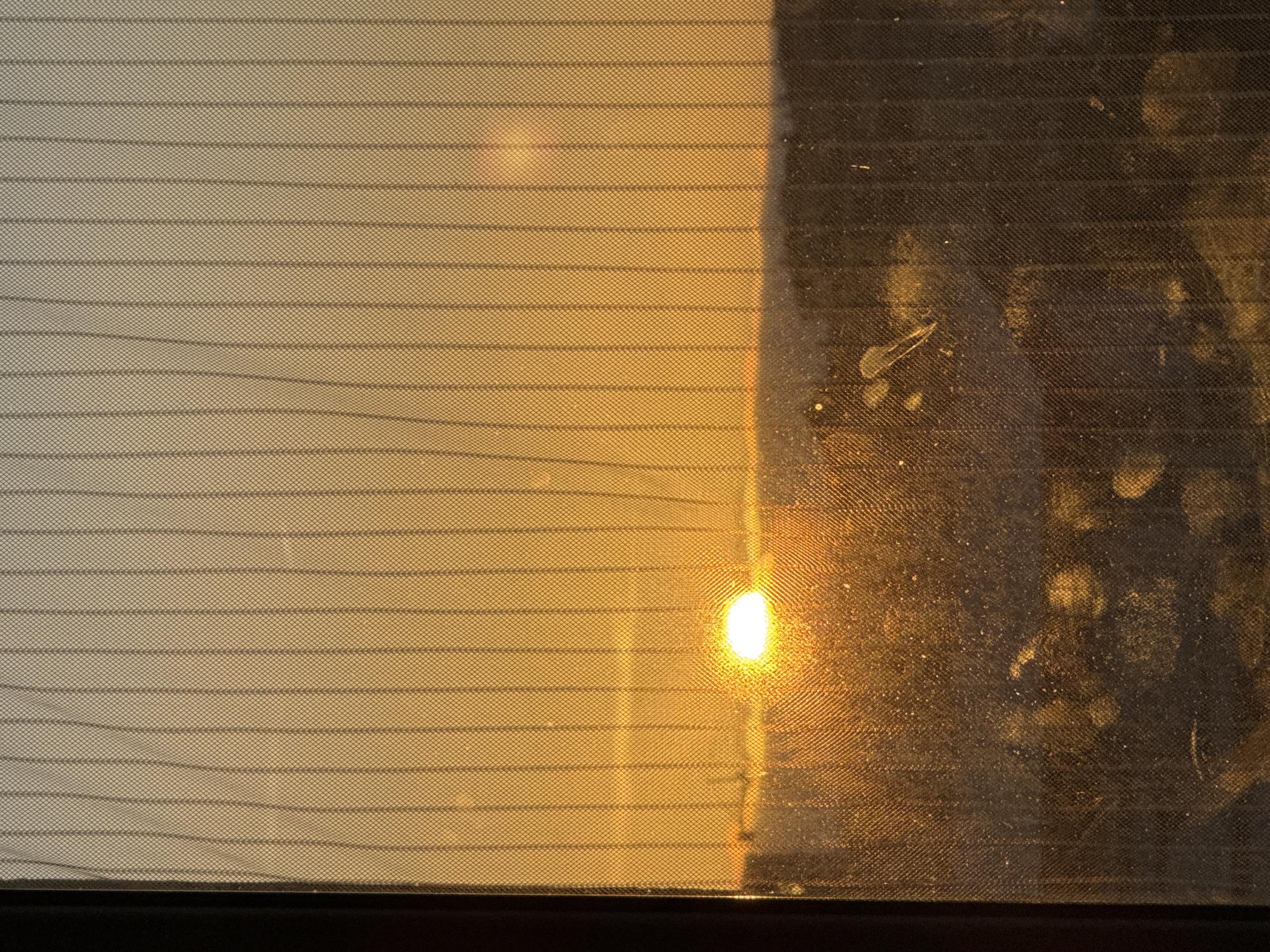 The image shows a hazy view through a rear car window with embedded heating elements, likely during a sunset or sunrise. The view is divided into two sections: the left portion shows a diffuse golden light passing through, while the right side is obscured by smudges and a film of dirt on the glass. A bright, circular light source, likely the sun, is visible through the center-right portion of the window. The bottom edge of the window is defined by a dark, presumably black, horizontal line. The overall scene gives the impression of looking at a landscape through a dirty window during a time when the sun is low in the sky. Figueira da Foz, Portugal is not visibly identifiable in the image.
