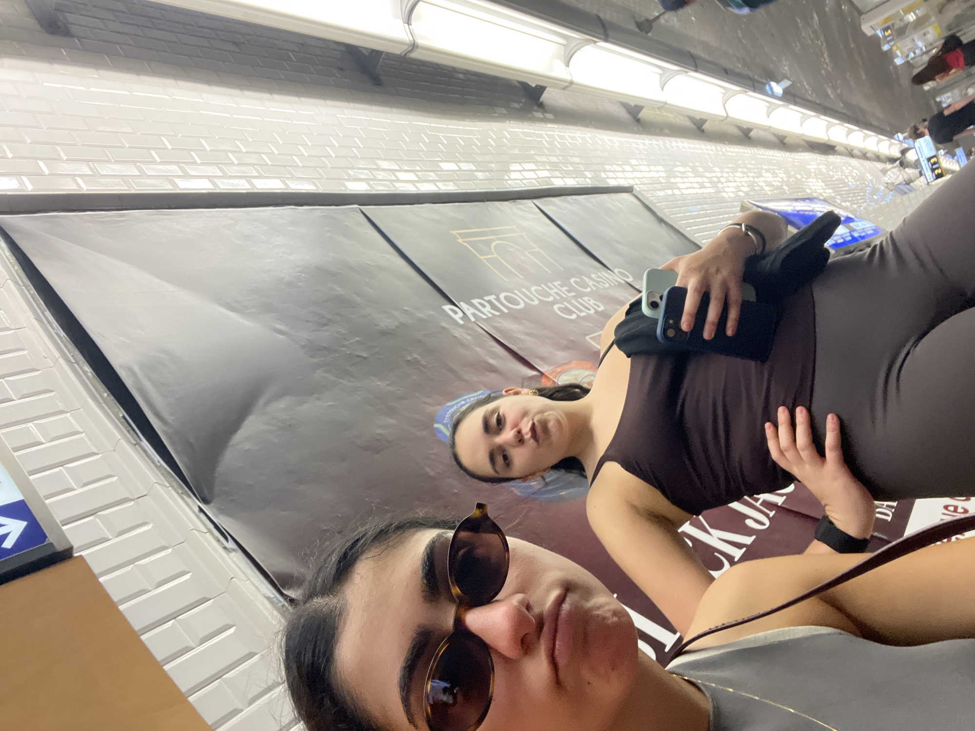 Two women are in a Paris Metro station, taking a selfie. The woman in the foreground wears sunglasses and a light-colored top. The other woman is behind her, wearing a brown top and gray leggings. A "Partouche Casino Club" advertisement is visible on the wall behind them. The wall is tiled with white subway tiles. Fluorescent lights illuminate the scene from the ceiling.