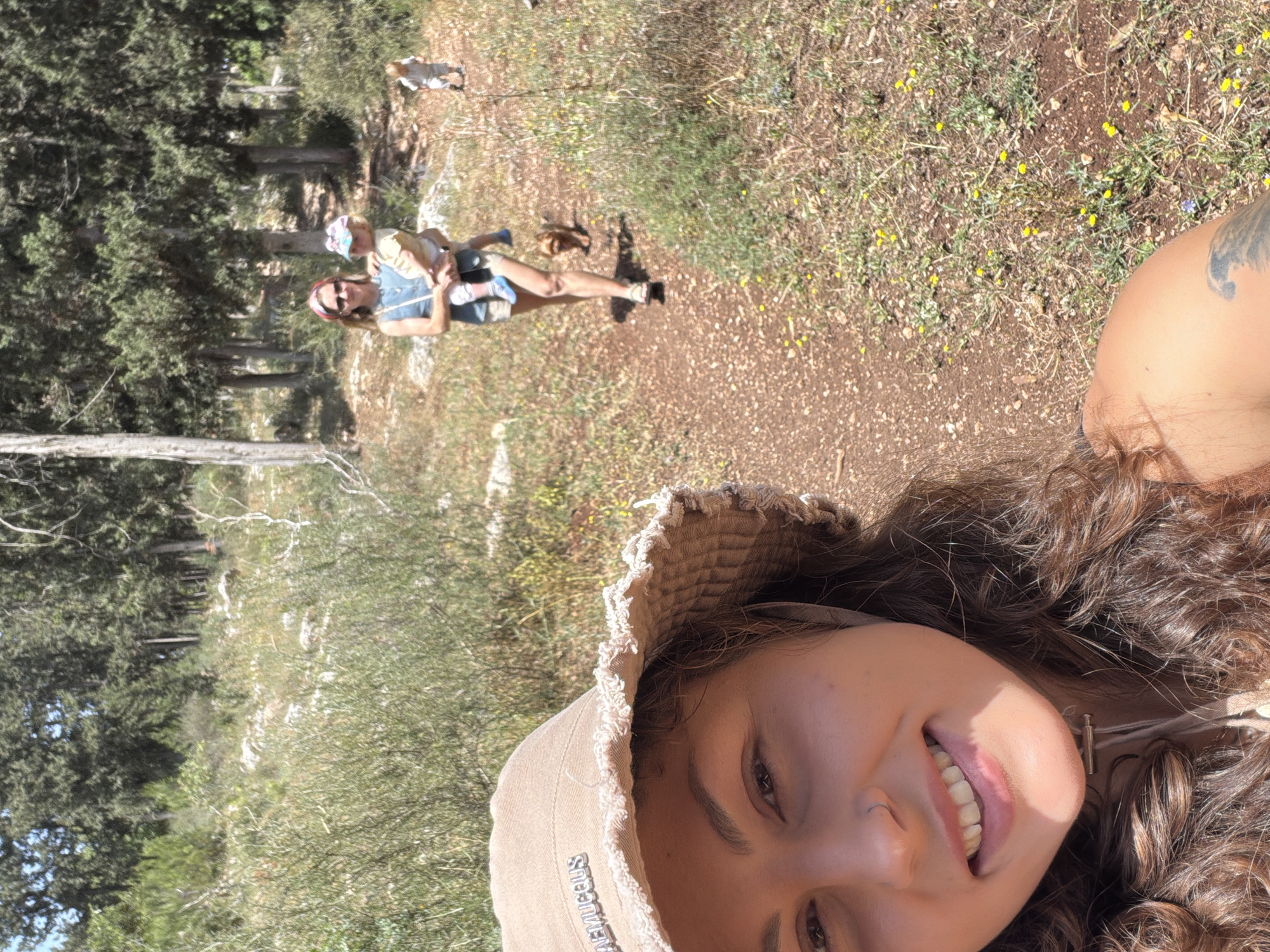 The image shows a smiling woman in a hat taking a selfie on a trail in what appears to be a natural area, possibly near the city of Zvulun Regional Council, Israel. In the background, another woman carries a child on the same path. The environment is characterized by trees, vegetation, and a dirt trail.