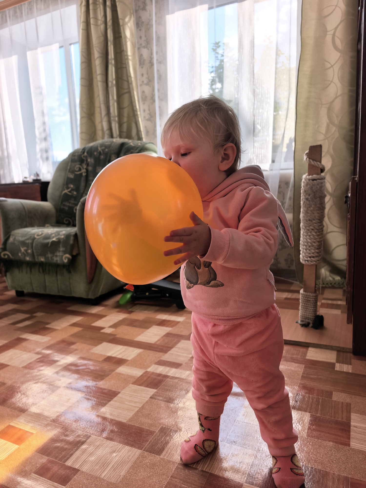 A toddler, dressed in a pink hooded sweatshirt with bunny ears and matching pants, attempts to inflate an orange balloon. The child's face is pressed against the balloon as they exhale. They are wearing pink socks decorated with fruit slices. In the background, a green armchair is visible, draped with a patterned blanket. To the right, a cat scratching post stands near a doorway. The scene appears to be indoors, on a floor with a checkered pattern of wood-look tiles. The lighting suggests it is daytime, with sunlight streaming through sheer curtains covering a window. The specific location of Sterlitamak, Russia, cannot be determined from the visual information present in the image.