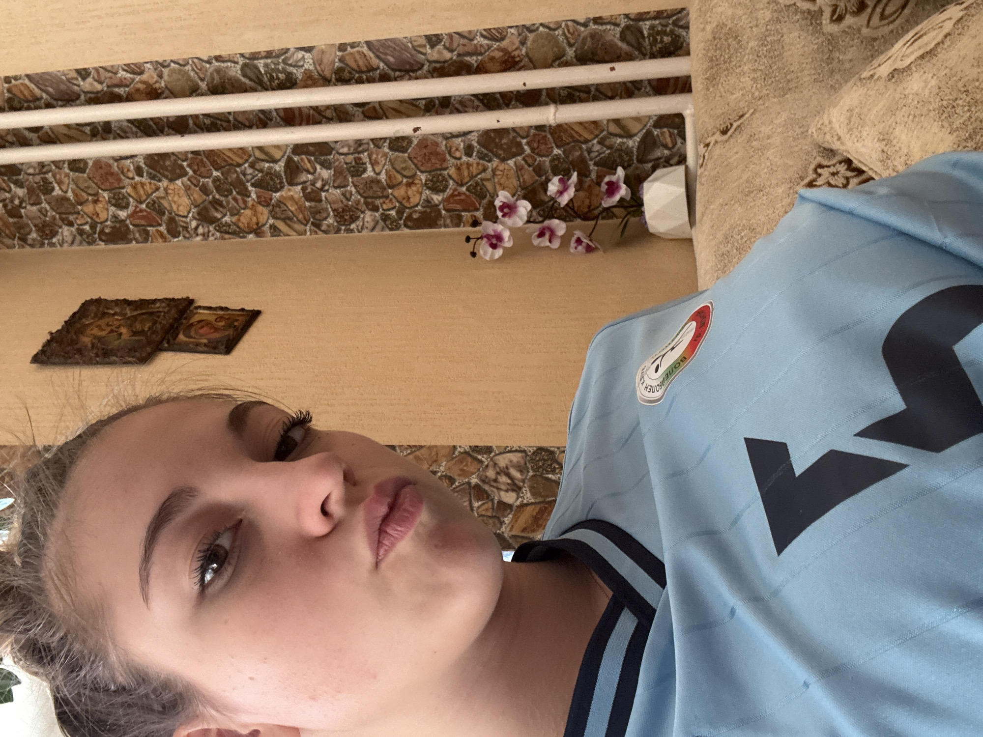 A woman is pictured indoors. She is wearing a light blue shirt with black stripes on the collar and a logo near her right shoulder. The background features a wall with a stone pattern design on the upper portion, with two framed pictures, and a faux floral arrangement. Based on the logo visible on the shirt, the photo was likely taken in Sofia, Bulgaria.