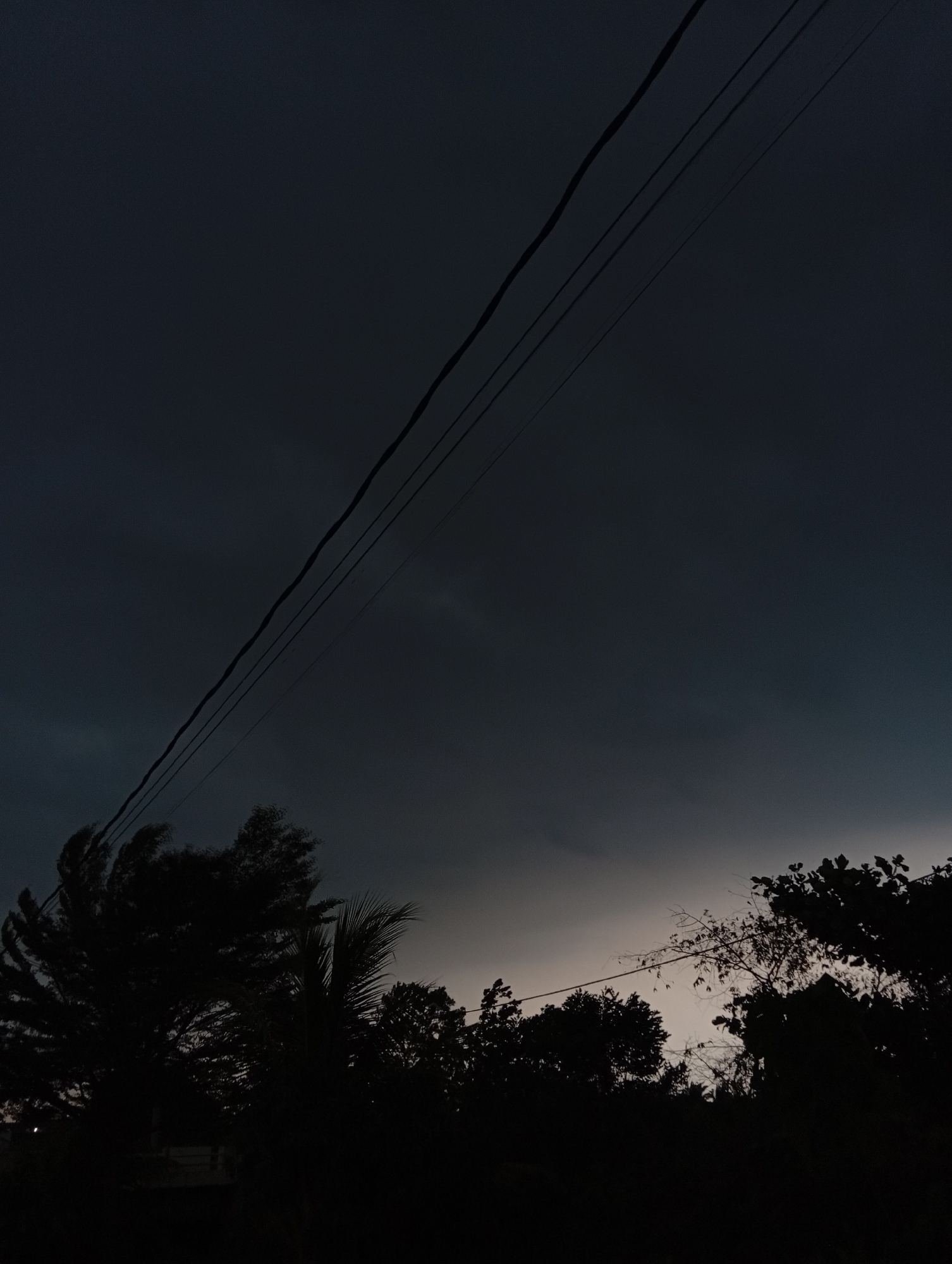 The image captures a dusky sky over Kualu, Indonesia, dominated by dark, brooding clouds that suggest an approaching storm or the twilight hours. Three dark power lines stretch diagonally across the upper portion of the frame, their taut lines contrasting with the organic shapes of the silhouetted trees that form a dense, dark band across the lower edge of the image. Among the trees, specific foliage types are not clearly discernible due to the low light conditions, though some appear wind-swept. The environment is devoid of any visible people or immediate action. The overall impression is one of impending weather change or the transition from day to night in a semi-urban or rural setting.