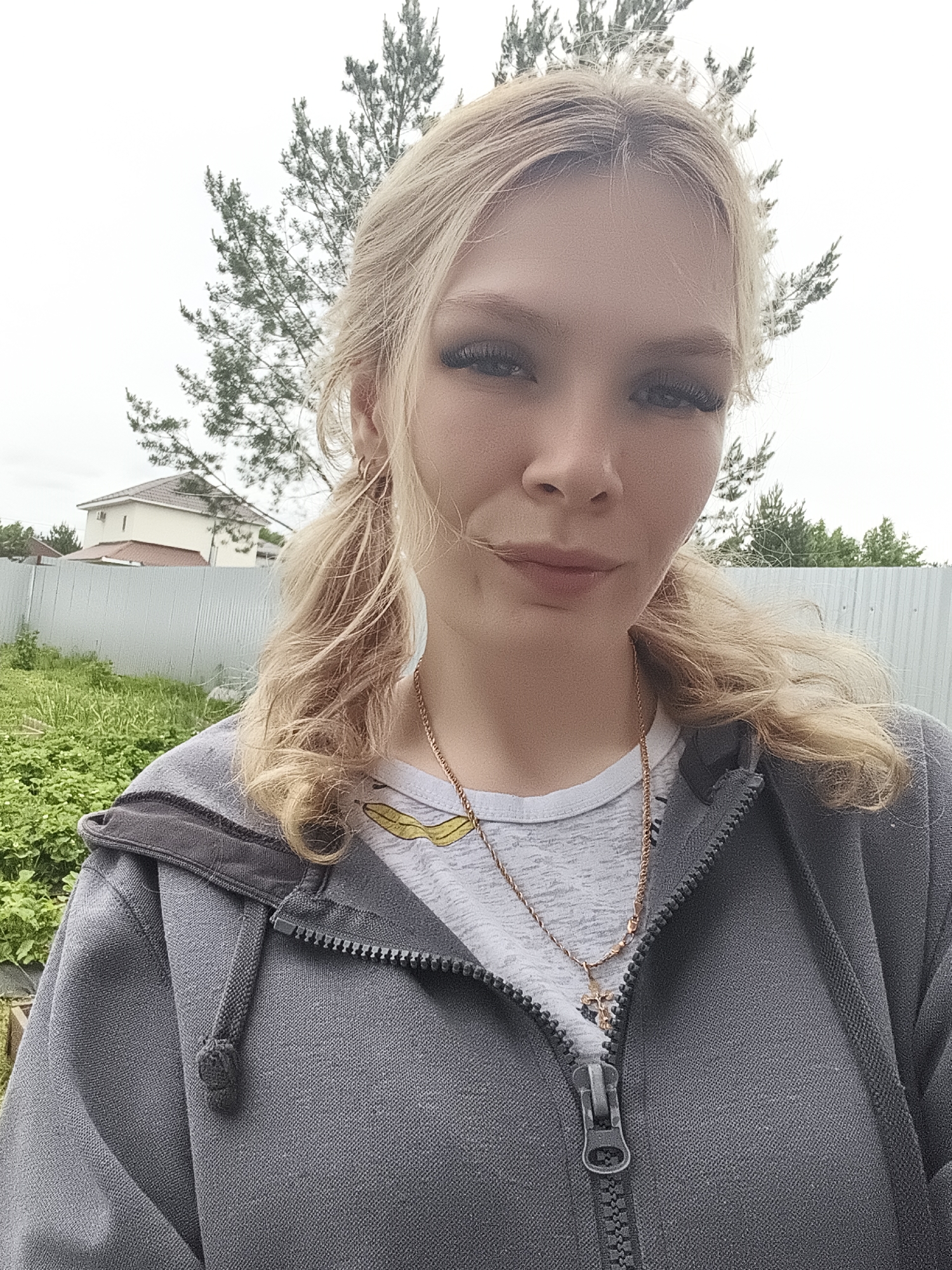 The image shows a young blonde woman with her hair in two pigtails, wearing a grey hooded jacket and a white t-shirt with a yellow graphic. She has on a gold necklace with a cross pendant. She is standing outdoors in what appears to be a suburban or rural setting, with a metal fence and a house in the background. Lush green vegetation is visible in the foreground and to the left of the woman. The sky is overcast. The specific location within Русский Вожой, Russia is not determinable from the image alone.