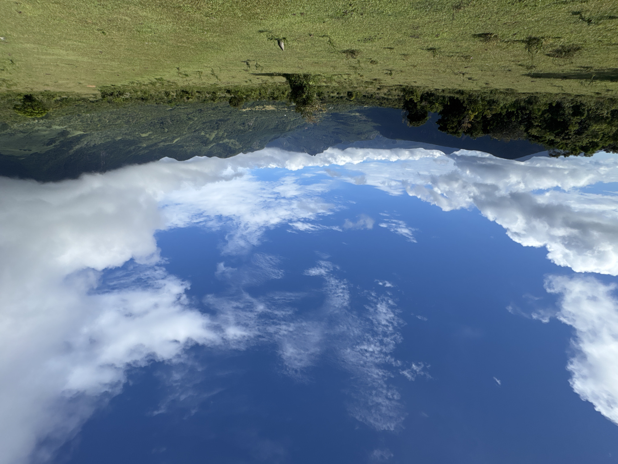 The image depicts an upside-down landscape, likely taken in Illukkumbura, Sri Lanka. The sky, predominantly a deep blue with scattered cirrus clouds and larger cumulus formations along the horizon, occupies the lower portion of the image. Above this horizon, a band of green, heavily forested mountains is visible, partially obscured by the clouds. The top section of the image showcases a grassy field with scattered trees. The image orientation gives the impression that the sky is a lake reflecting the landscape.