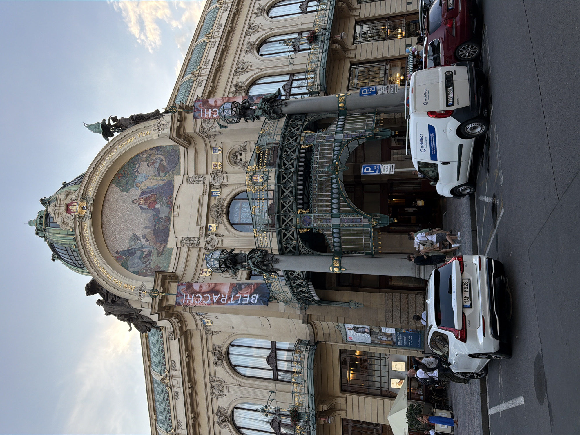 The image shows the Municipal House (Obecní dům) in Prague, Czechia, an Art Nouveau building located at Náměstí Republiky 5. In the foreground, a white Volvo V60 wagon, license plate 1AM F928, is parked. To its right, a white Mobiltel van, license plate 7AS 5002, is also parked. Several people are visible walking and standing on the sidewalk in front of the building. The Municipal House features a grand facade with elaborate sculptures, mosaics, and ornate balconies. Two vertical banners with portraits are hanging from the building. The sky above is partly cloudy.