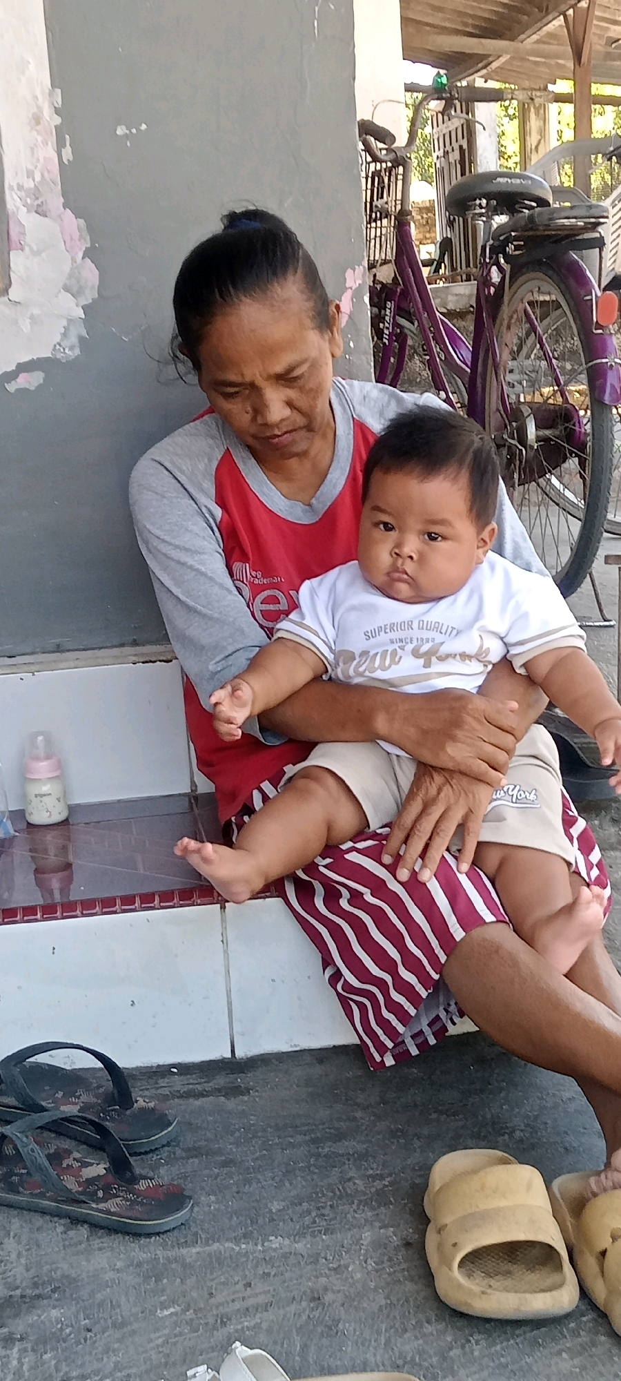 A woman in a red and grey shirt is holding a baby wearing a white t-shirt with "SUPERIOR QUALITY NEW YORK" printed on it. The woman is seated on a ledge with white tiled steps leading down. Next to her on the ledge is a baby bottle. The background shows a purple bicycle parked next to a structure with a wooden roof. The scene appears to be outdoors, possibly in the city of Juwiring, Indonesia, with a concrete floor in the foreground where a pair of flip-flops and some other footwear are visible.