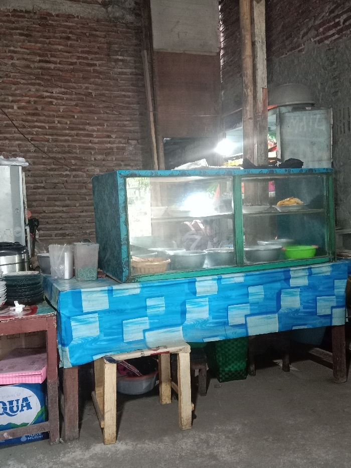 An indoor scene shows a modest food stall. A blue and white tablecloth-covered table holds a glass display case filled with dishes. To the left, a stack of plates sits on a red-trimmed table with a blue water bottle beneath. A small wooden stool is positioned in front of the main table. A brick wall is in the background.