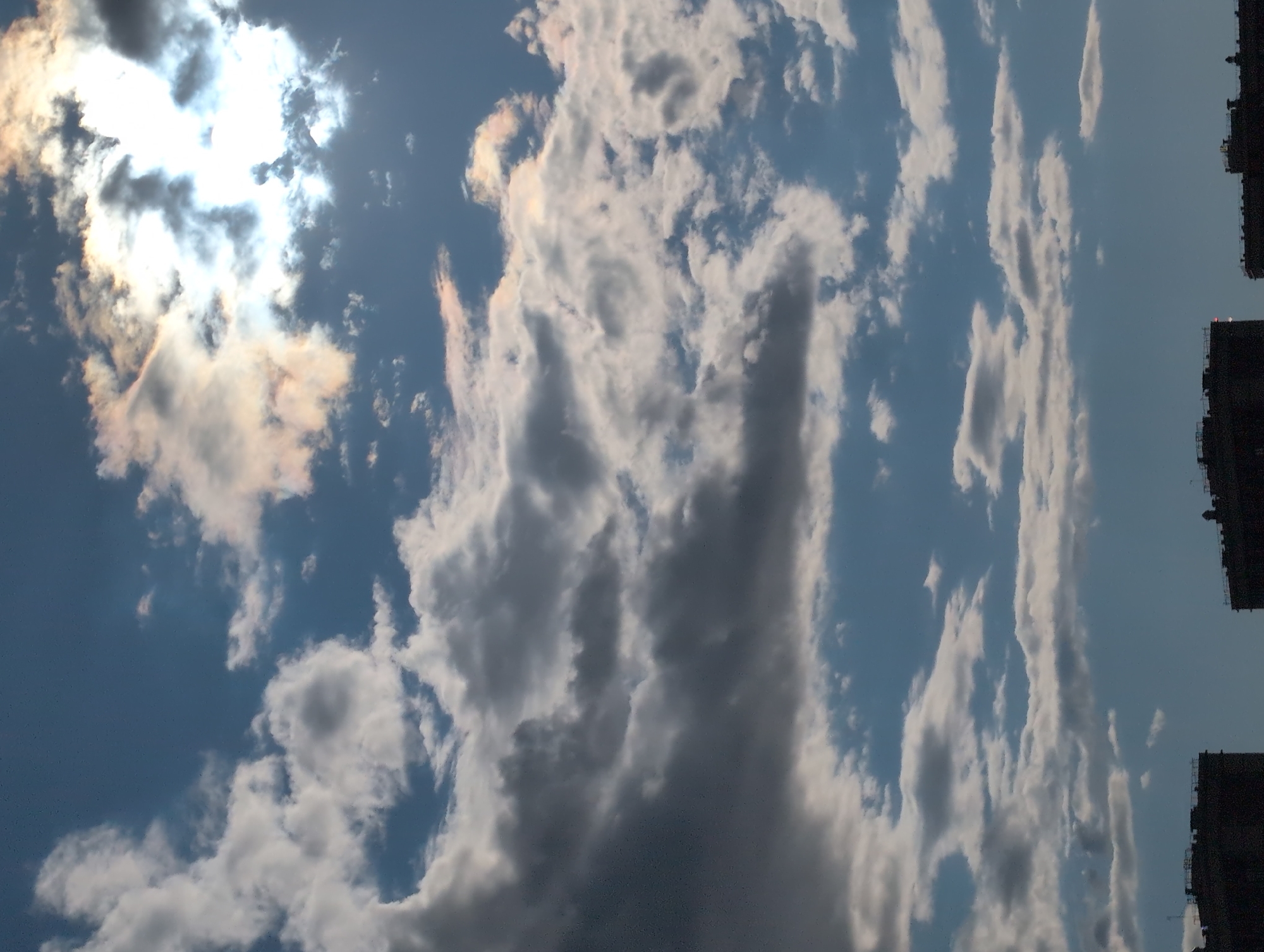 The image is a vertical, low-angle shot of the sky above Saint Petersburg, Russia, with a portion of buildings visible on the right edge. The sun is partially obscured by thick, white and gray clouds, creating a dramatic and bright halo effect. Wispy, lighter clouds are scattered across the blue sky, and some of the clouds exhibit iridescent colors, suggesting atmospheric phenomena like iridescence. The presence of buildings indicates an urban setting. The overall lighting suggests it is daytime, possibly late afternoon or early evening given the warm tones in some of the clouds and the angle of the sun. There is no visible text. The scene evokes a sense of wonder and natural beauty.