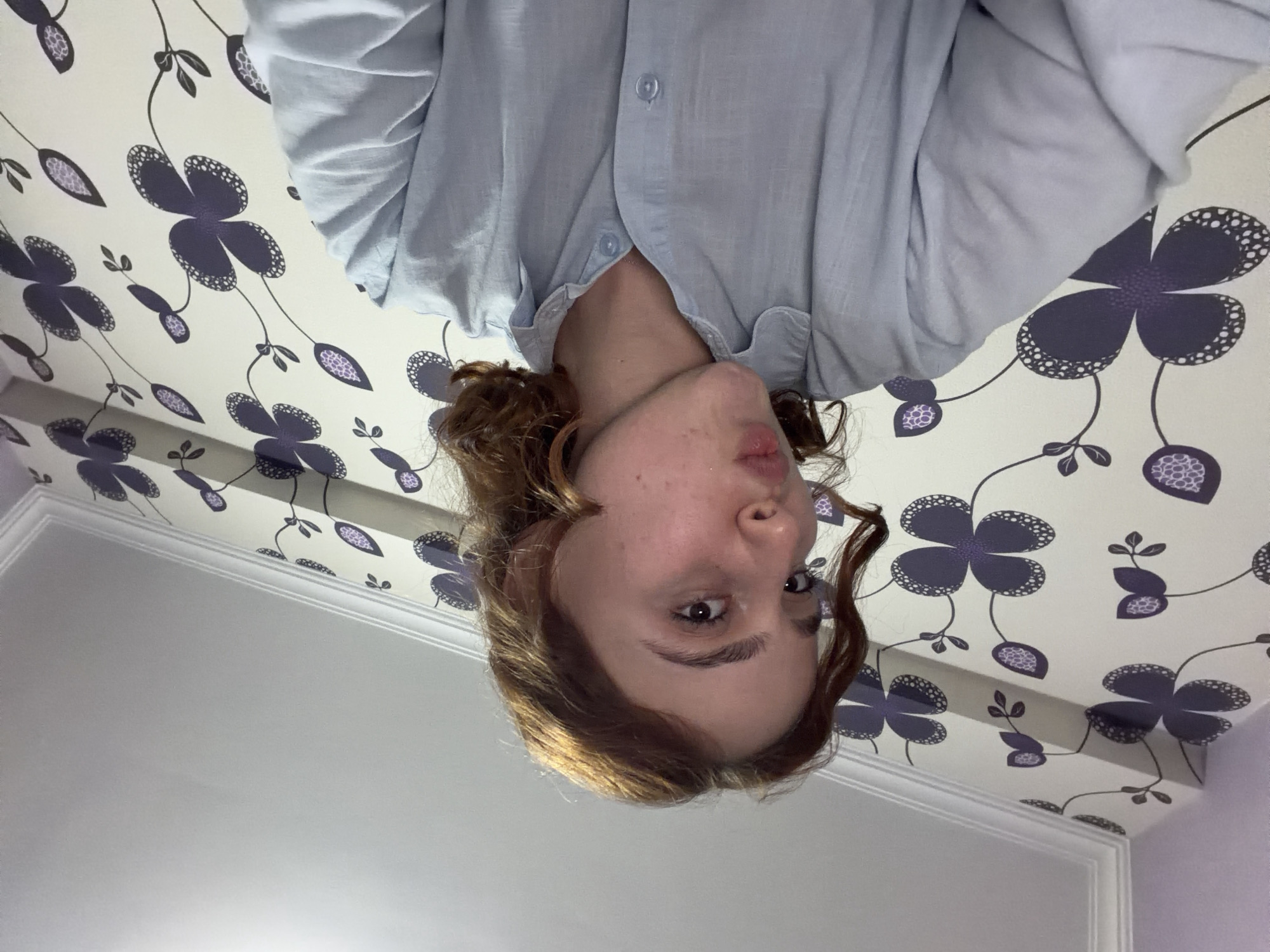 A person is positioned upside-down, facing the camera. They have light brown hair, fair skin, and are wearing a light blue button-down shirt. The background consists of a white wall with a floral wallpaper pattern of purple flowers. A portion of a white ceiling and ornate molding are also visible. The image's orientation suggests the person might be lying on their back or the camera was rotated. There is no visible information to indicate the location is Tbilisi, Georgia.