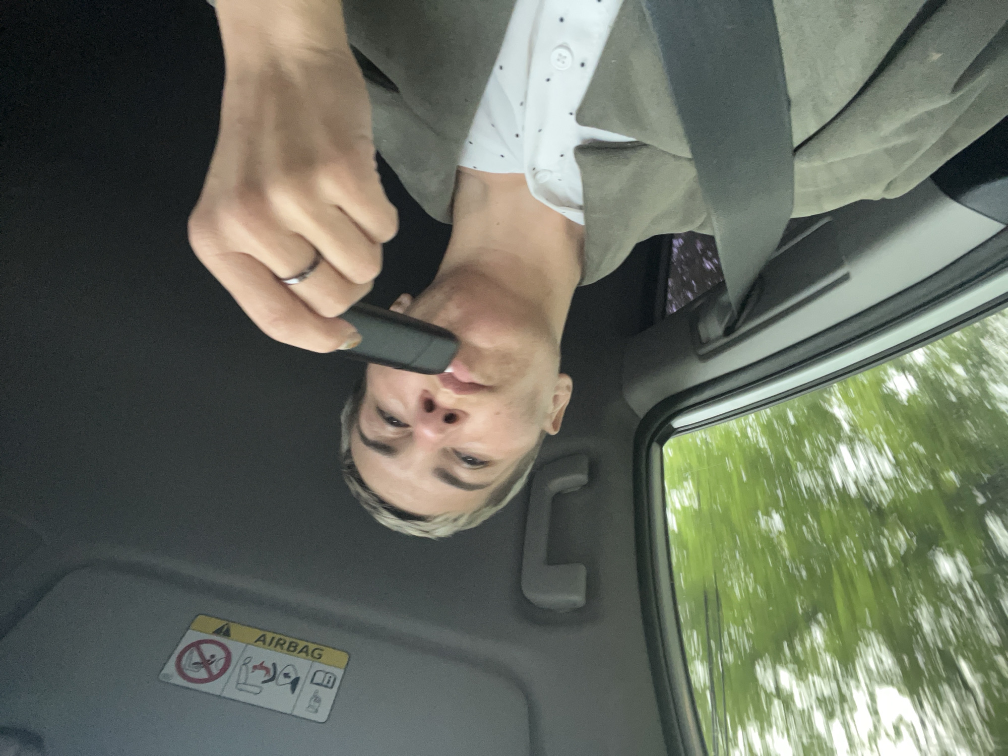 A photograph shows a person in the front passenger seat of a moving vehicle. The person is wearing a light-colored, button-down shirt with dark spots, and a khaki jacket. A seatbelt is fastened across their body. The person is holding a black device near their face, with a ring visible on one of their fingers. The window shows green trees in motion, indicating the vehicle is moving. An "AIRBAG" warning label is visible on the vehicle's sun visor. The photo is taken at an angle, looking up at the person and the vehicle's interior. The location is not discernible.