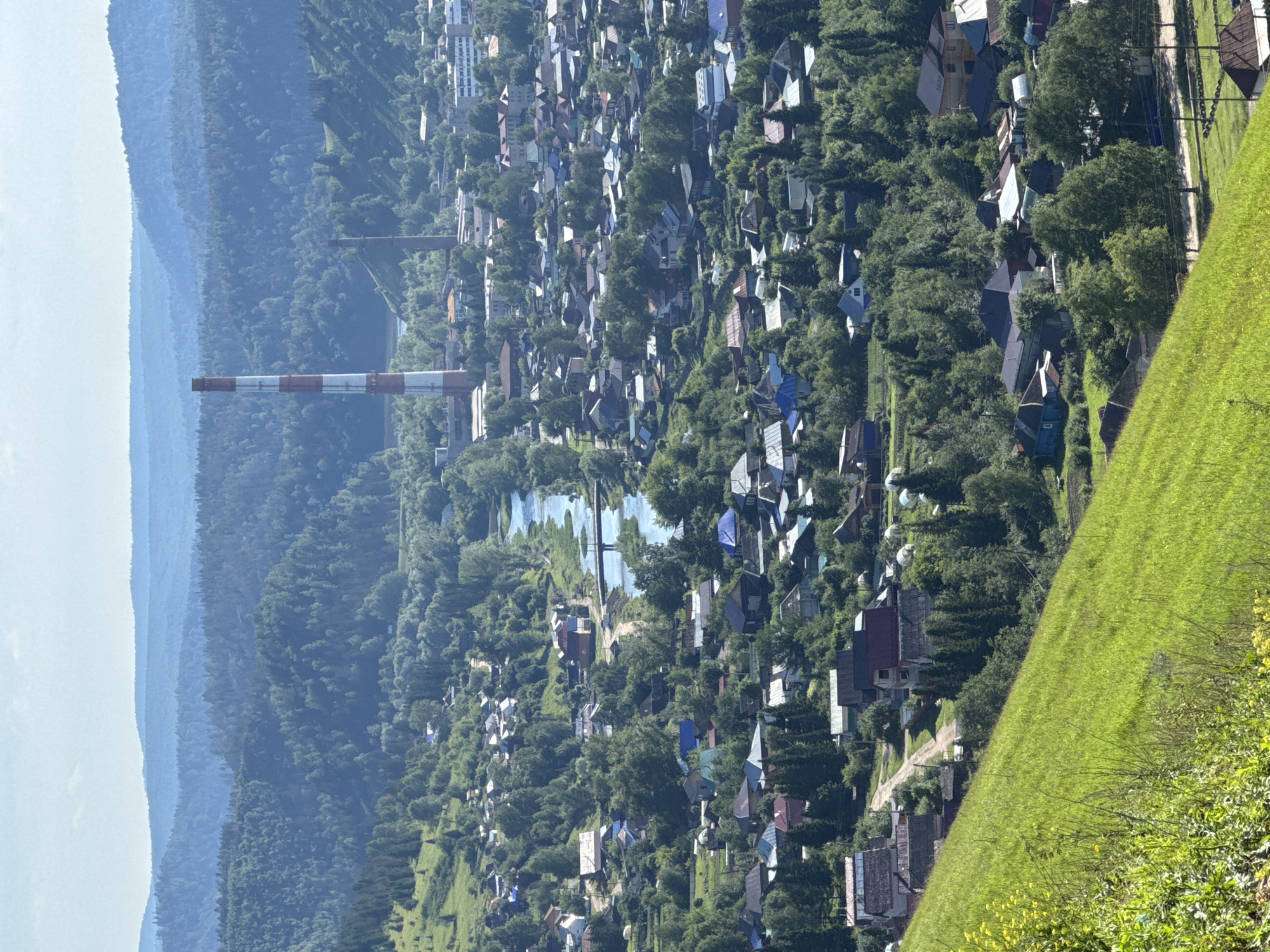 This is a high-angle, medium-shot photograph of the city of Sim, Russia, on a sunny day. The foreground is dominated by a steep, vibrant green grassy slope dotted with yellow wildflowers on the left. The middle ground reveals a dense residential area nestled within a valley, characterized by numerous houses with diverse roofing materials and colors. A river snakes through the town, crossed by a bridge in the middle ground. In the background, a tall, striped industrial smokestack (red and white) stands prominently, with another smaller smokestack visible to its right. Beyond the town, layered, tree-covered hills recede into the distance, fading into a hazy blue horizon under a light blue sky. The overall scene depicts a settlement integrated with its natural, hilly surroundings.