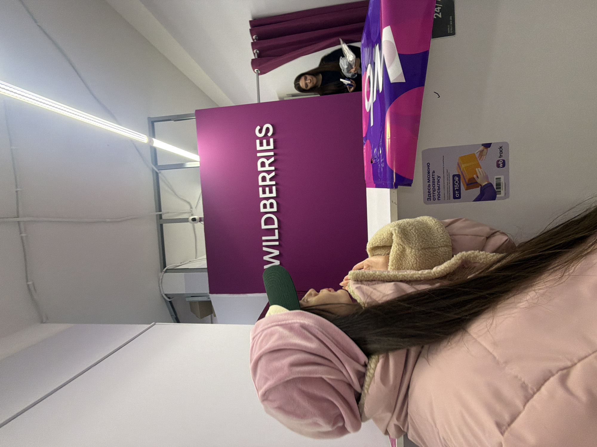 The image displays an indoor scene within a Wildberries pick-up point. While Wildberries points are prevalent in Saint Petersburg, Russia, the precise address within the city cannot be ascertained from the visual information provided.
In the foreground, positioned to the left, a female subject is captured from her right profile, encompassing her head and upper torso. She wears a light pink puffer jacket featuring a cream-colored fleece-lined collar. Her head is covered by a voluminous light pink velour material, worn over a dark green baseball cap with a beige underside brim. Her long, dark brown hair extends down her back. Her hands are clasped near her chin, and she is gazing towards the right, appearing to be waiting or observing.
A light-colored counter spans the middle ground of the image. On the right side of this counter, a partially flattened purple package, bearing the white "wb" logo consistent with Wildberries branding, is visible. Adjacent to it on the counter surface is a white sticker. The sticker displays Russian text: "Здесь можно отправить посылку" (Here you can send a parcel) and "ОТ 150Р" (FROM 150 RUB), along with an illustration of hands exchanging a package, a "wb track" logo, and a QR code. A partially visible sign to its right indicates "24/7" service.
Behind the counter, a prominent purple wall segment is centered, featuring the word "WILDBERRIES" in raised white letters, identifying the brand. To the right of this purple wall, a dark purple curtain hangs from a rail, partially obscuring a second female subject. This individual, possessing dark hair and wearing a dark long-sleeved top, is holding a white, somewhat reflective object in her left hand and another object in her right. She appears to be looking forward, possibly towards the foreground.
The environment consists of plain white walls and ceiling, illuminated by multiple linear fluorescent light fixtures. A small, white dome security camera is mounted high on the upper right wall area, and exposed white conduits or cables are visible on the ceiling.