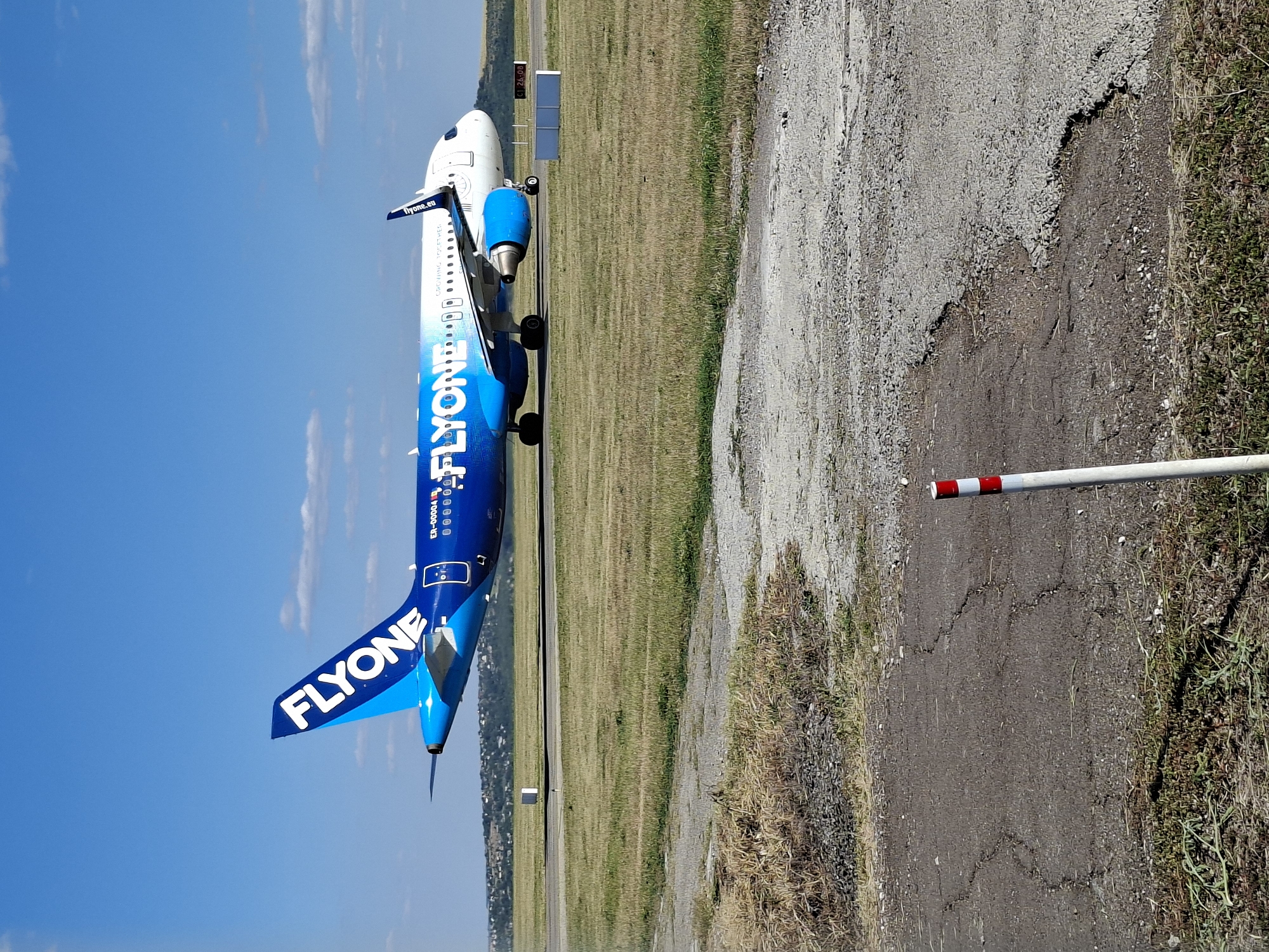 An overhead shot shows a bright blue airplane with "FLYONE" written on its side on a runway at Chișinău International Airport, Moldova. The airplane is white and blue with blue engines. The tail of the airplane is blue with "FLYONE" written in white letters. The plane is on a paved runway with grass on either side. The sky is a clear blue with a few white clouds. The airport is surrounded by rolling hills with trees and buildings visible in the distance. The image is taken from a low angle, looking up at the airplane. A red and white striped pole is in the foreground of the image.