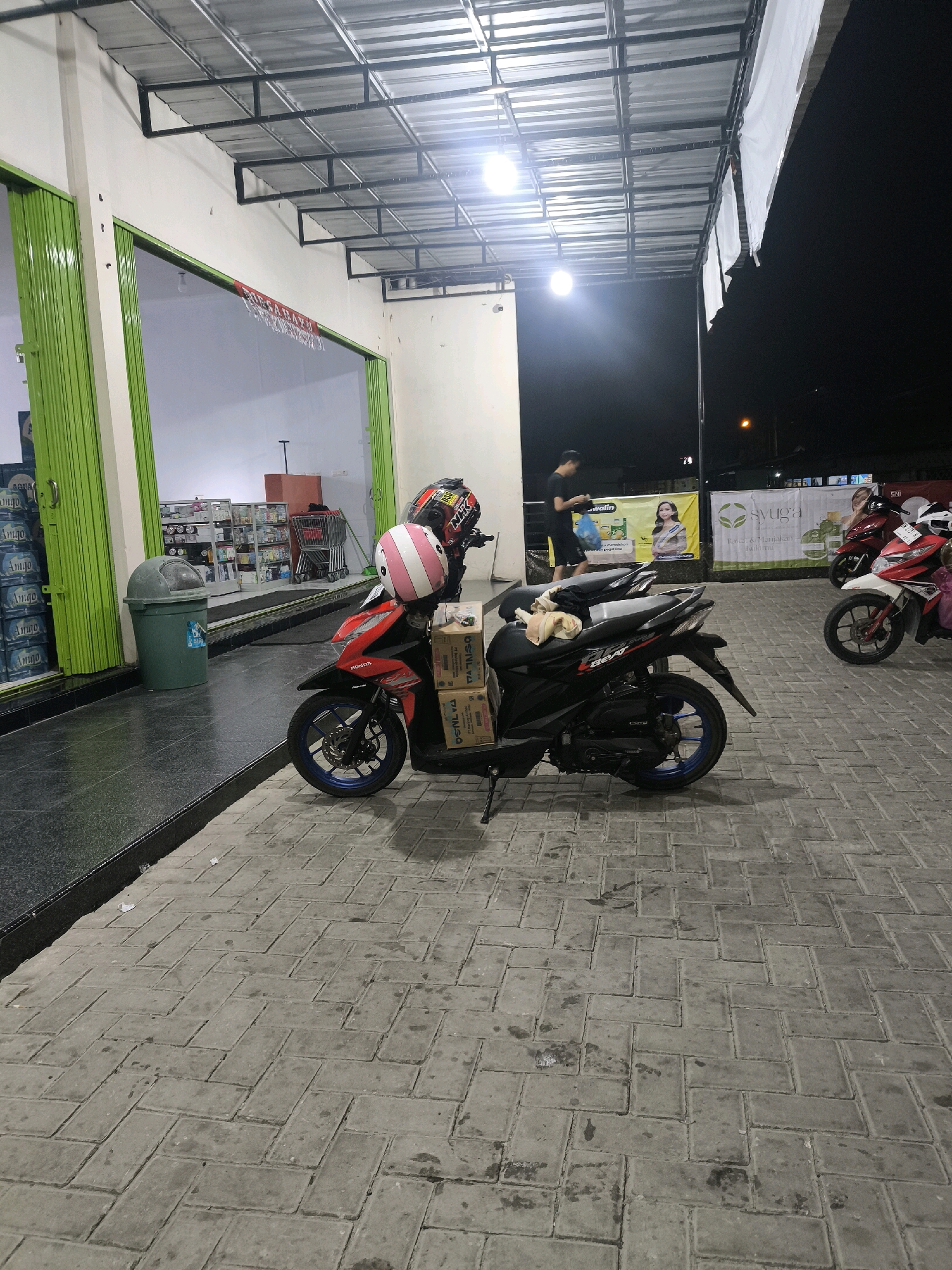 A motorcycle is parked on a brick sidewalk in front of a store. The motorcycle is black and red and has two boxes strapped to the back. There is a helmet on the seat of the motorcycle. In the background, there are shelves of products inside the store, and a shopping cart is visible. A person is standing near the entrance of the store, and another person is walking away from the store. The scene appears to be at night, as indicated by the lighting and the dark sky. The location is identified as Pilolahuwa, Indonesia.